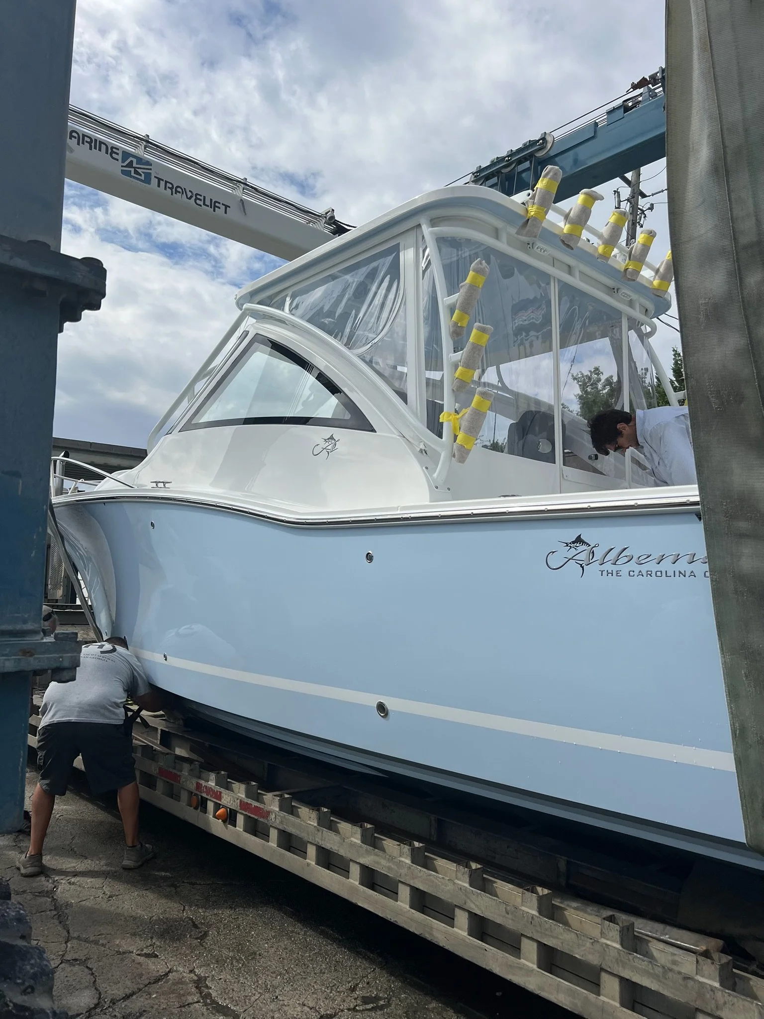 A large boat is being transported on a flatbed trailer with two men working on it. The boat has a white superstructure with clear windows and black detailing, and is secured with yellow tape on several parts. There is a blue crane with the marking 'T