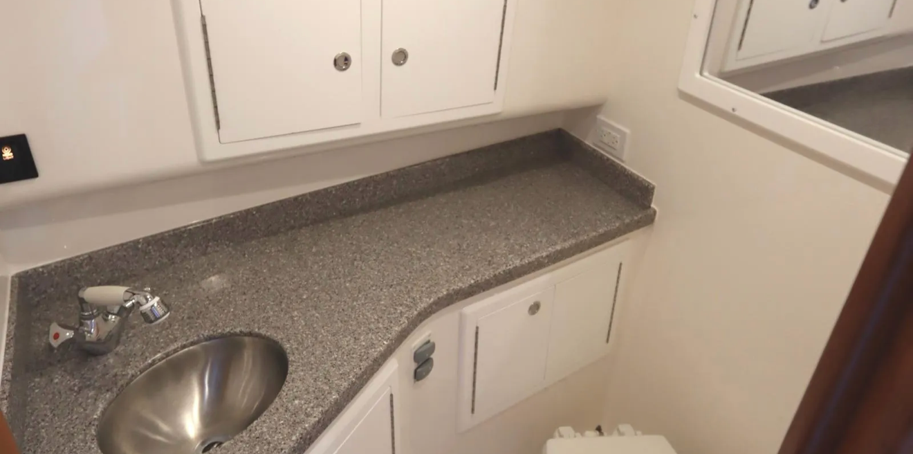 Empty kitchenette with a gray speckled countertop, small round stainless steel sink, white cabinets above and below, a window, and electrical outlets.