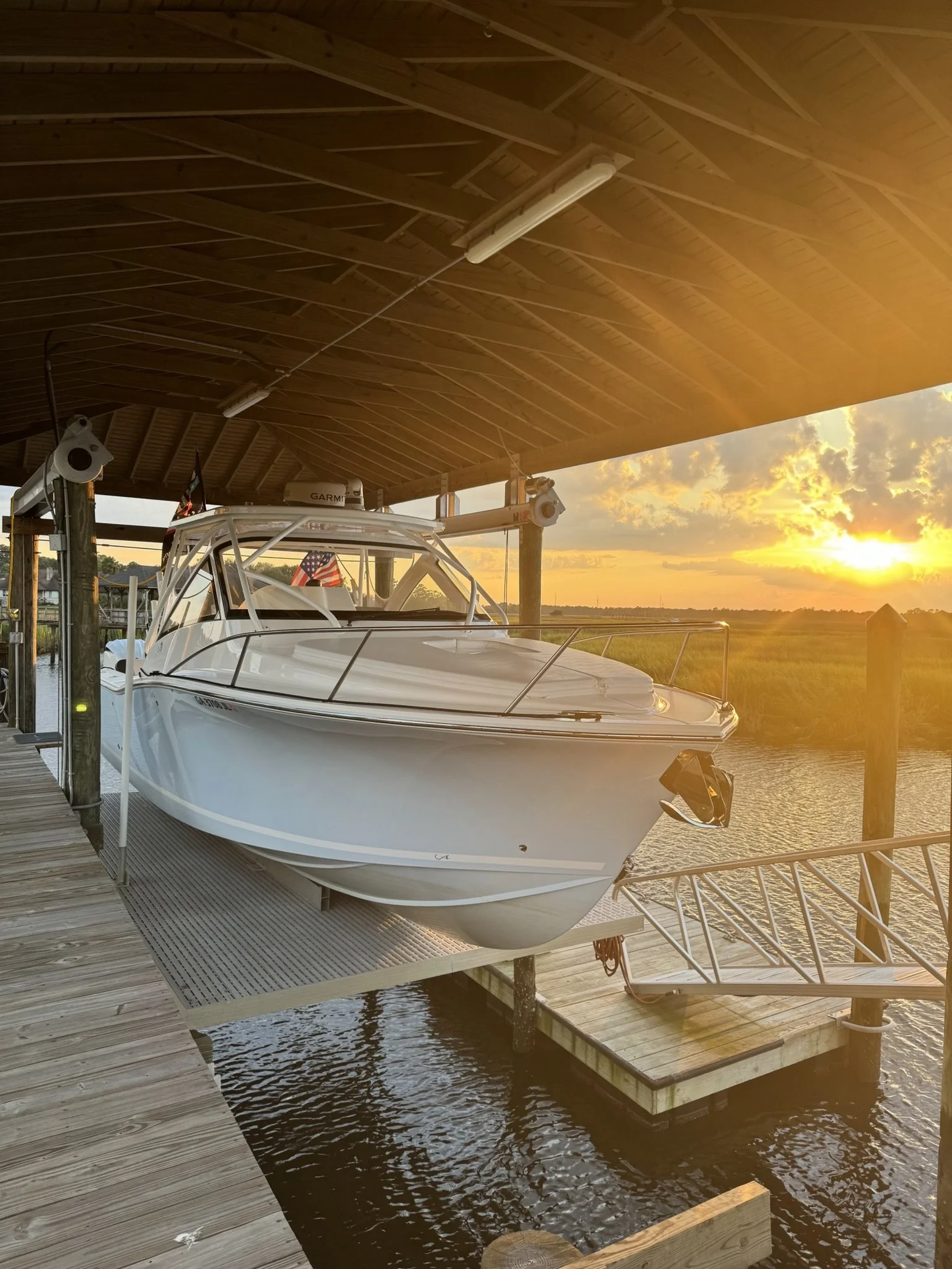 A white yacht docked at a wooden pier under a covered boathouse during sunset, with a calm waterway and grassy field in the background.