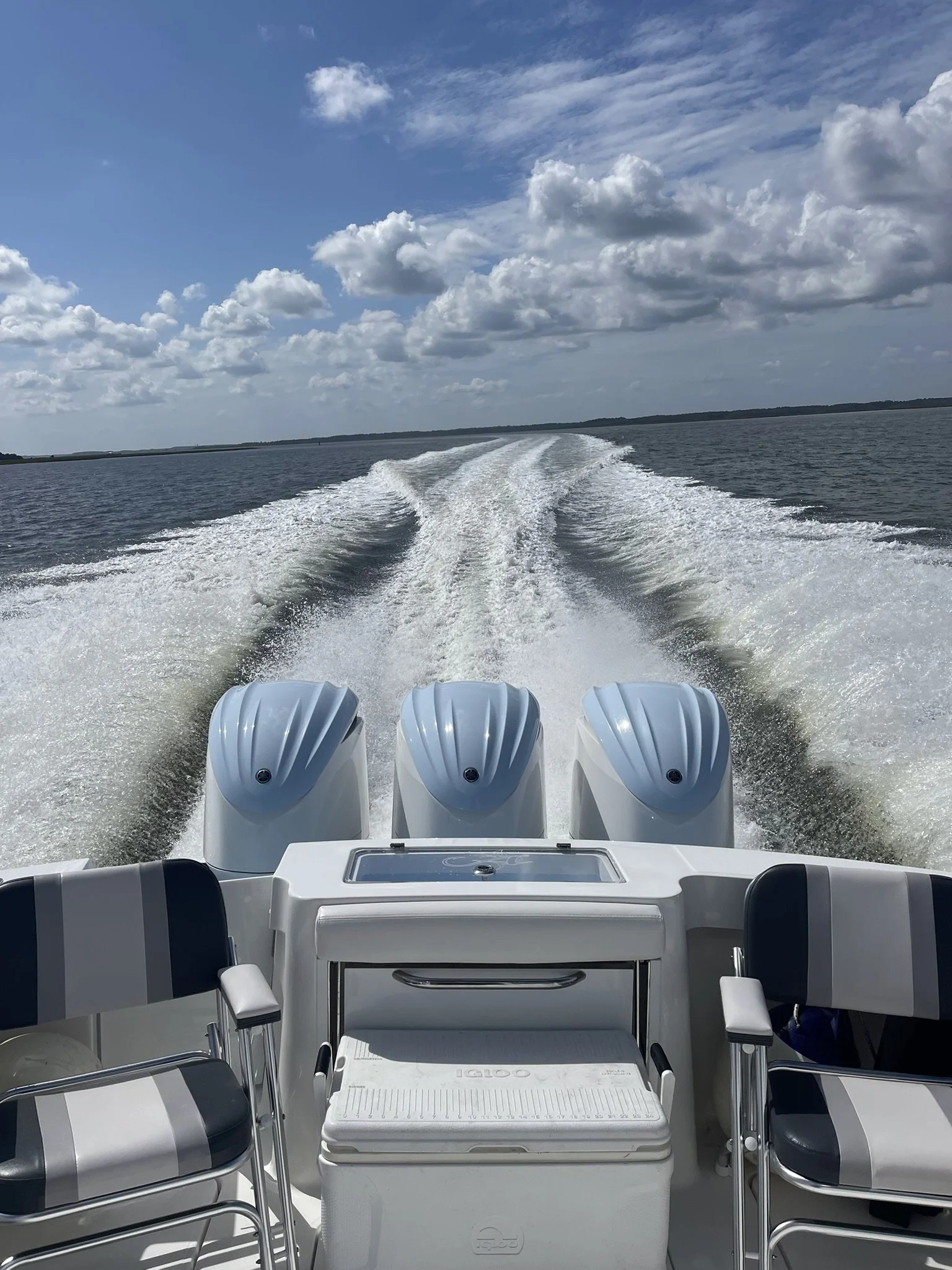 View from the back of a boat showing three outboard motors, a small table or cooler, two chairs, and water trail in the water behind the boat with blue sky and clouds above.