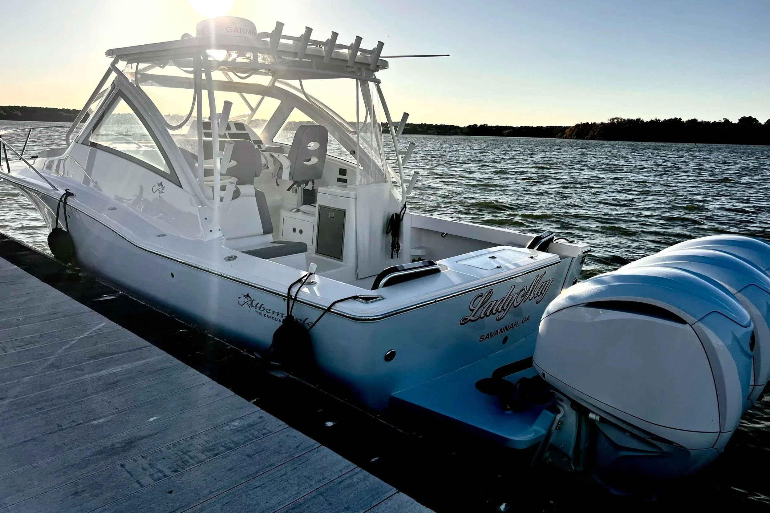 White motorboat docked at a marina with a body of water and trees in the background during sunset.