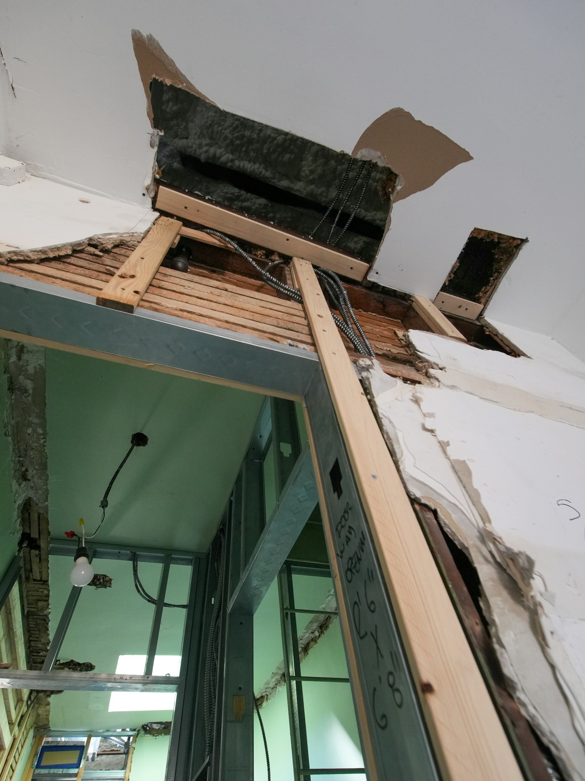 Construction site showing exposed wall with broken drywall, electrical wiring, and framing. Inside space with unfinished ceiling, green wall, and hanging light bulb.