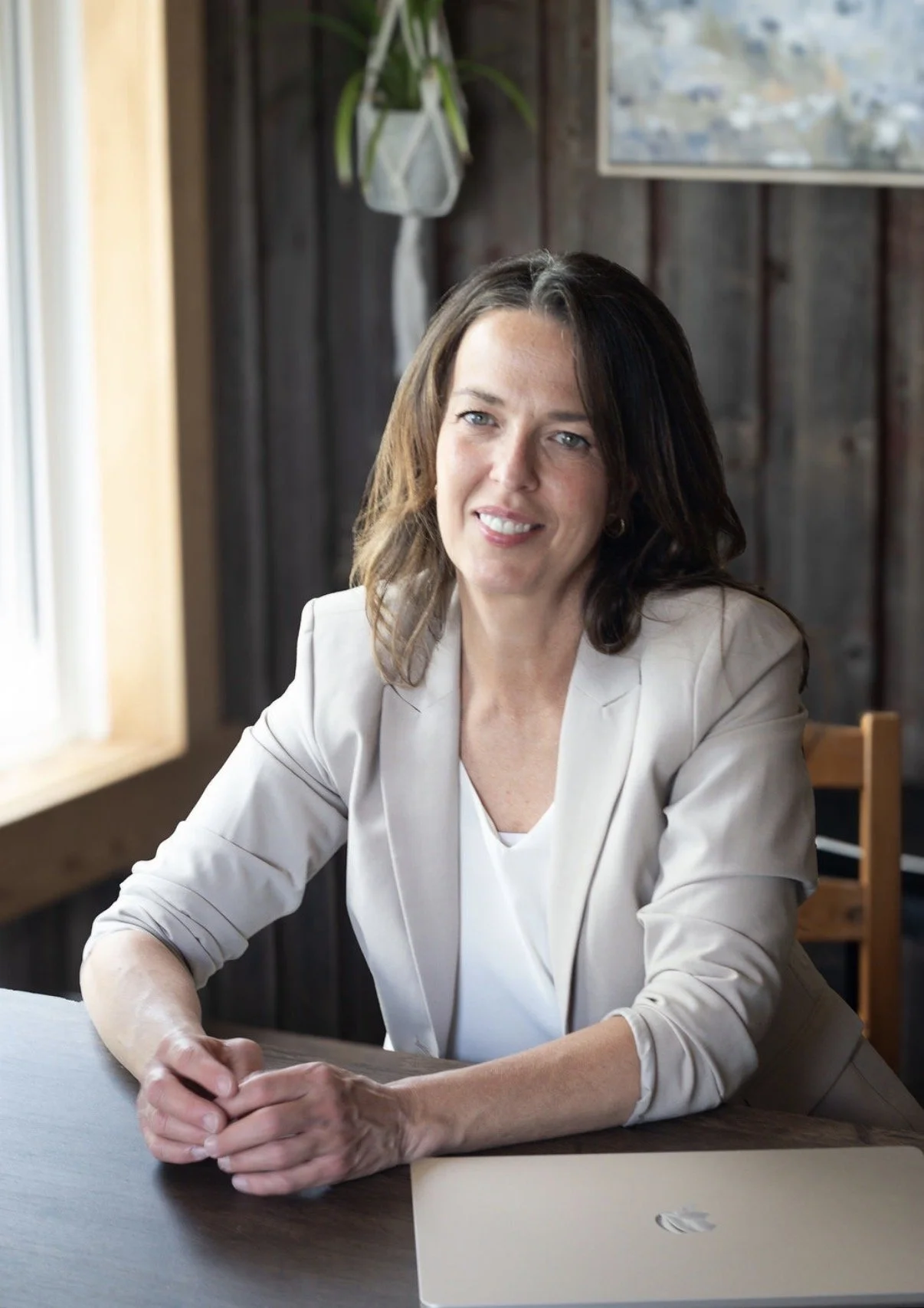 A woman with brown hair and blue eyes sitting at a table, wearing a cream-colored blazer over a white shirt, smiling and looking at the camera. A laptop is on the table in front of her, with a paper beside it. The background features wooden walls, a window on the left, and a plant hanging on the wall.