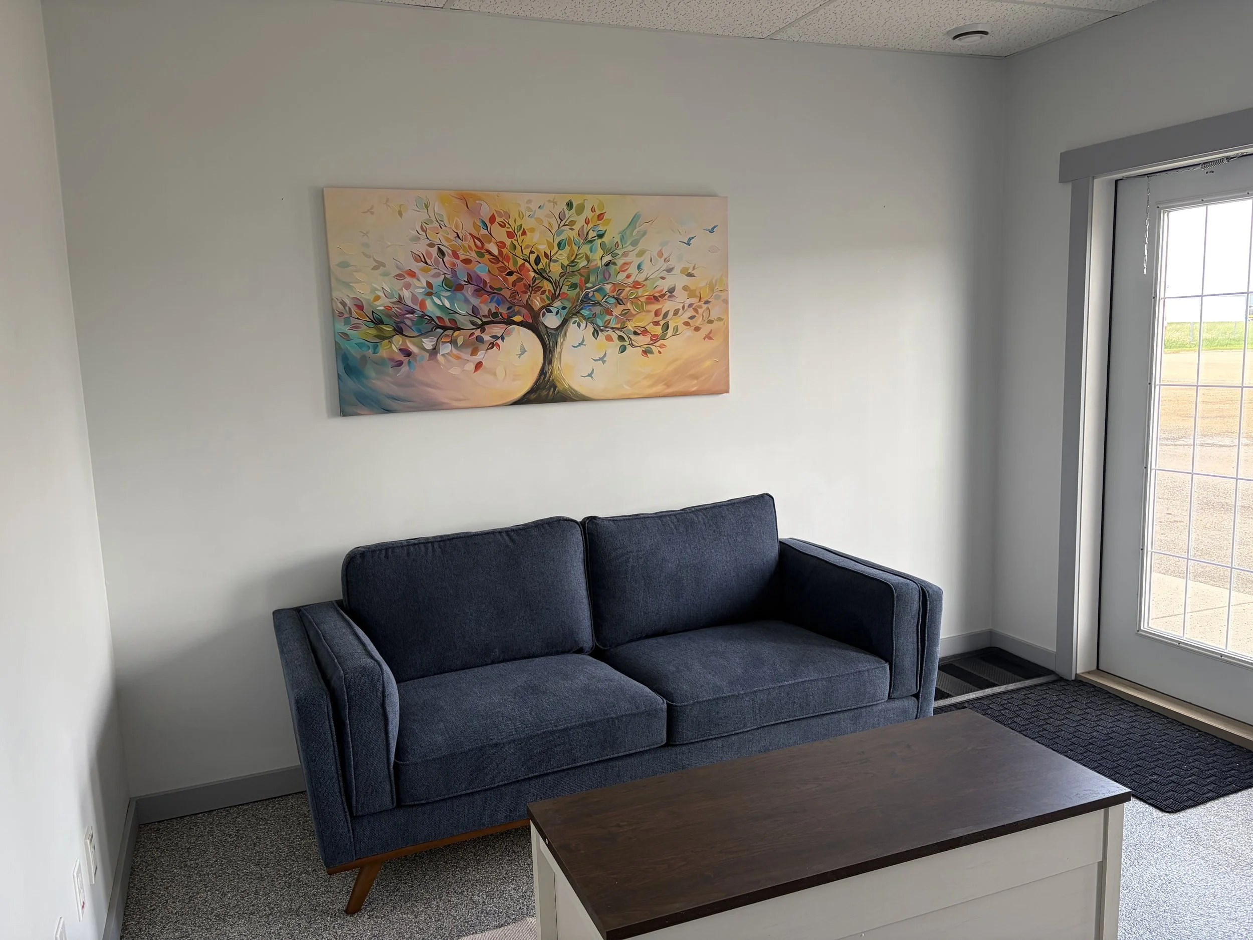A small, dark blue sofa with wooden legs is positioned against a white wall. A colorful painting of a tree with multicolored leaves is hanging above the sofa. To the right, a glass door leads outside, revealing a garden or field. The room has a dark wooden coffee table in front of the sofa.