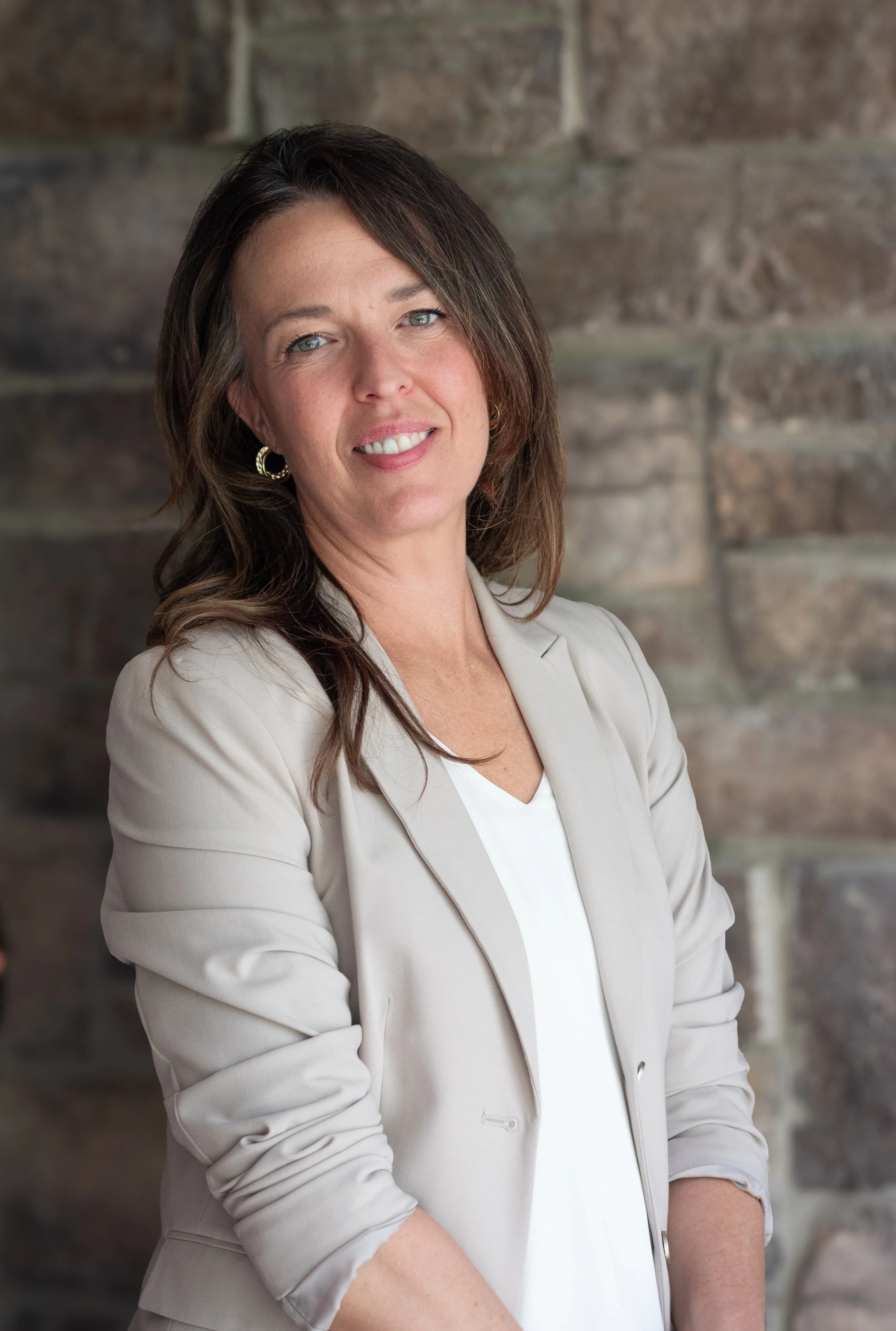 A woman with brown hair, green eyes, wearing a beige blazer and earrings, smiling, standing in front of a stone wall.