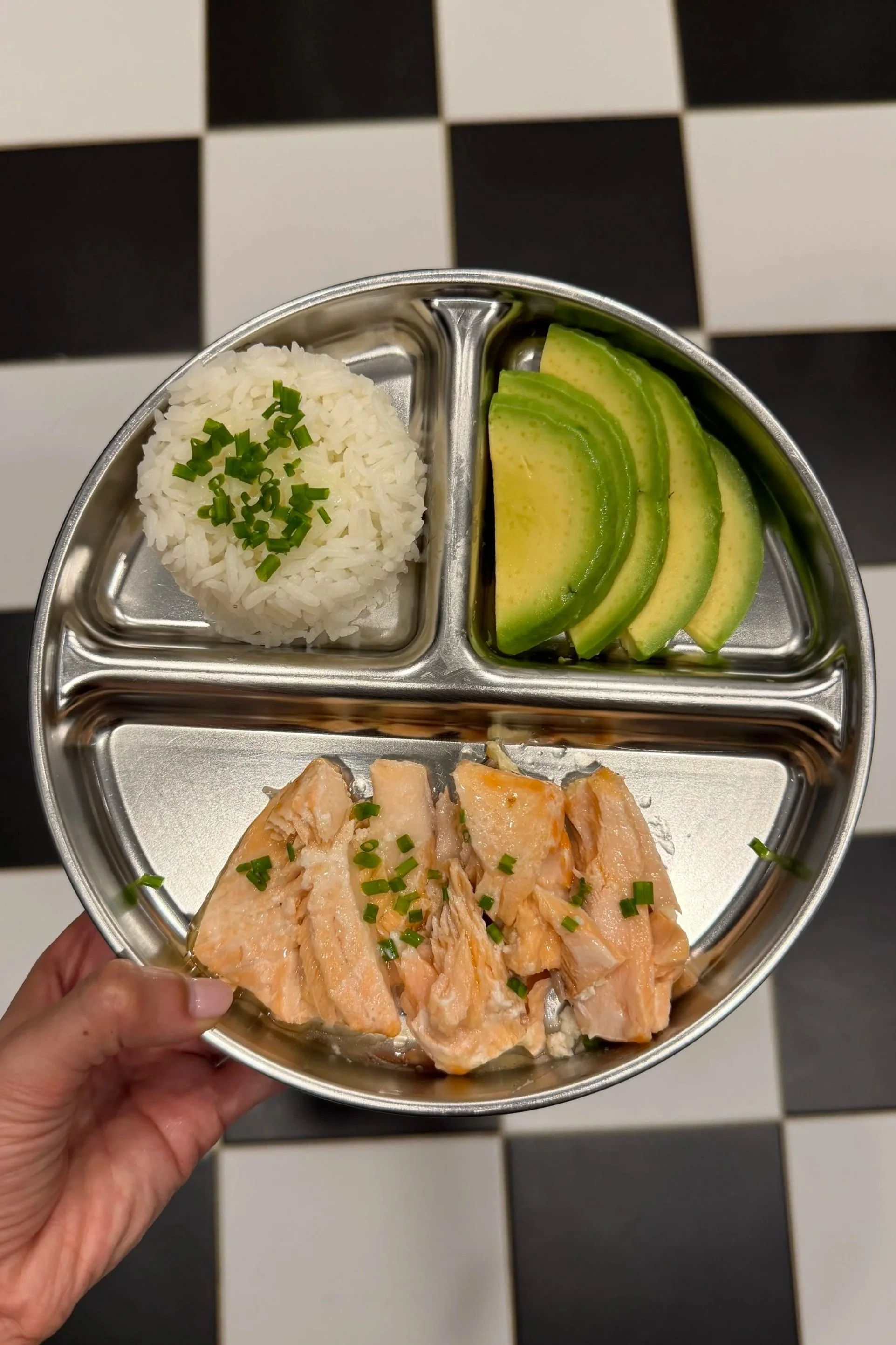 A stainless steel divided tray containing three sections: one with cooked white rice garnished with chopped green onions, another with sliced avocado, and the third with pieces of cooked salmon also garnished with chopped green onions. The tray is held by a person's hand over a black and white checkered floor.