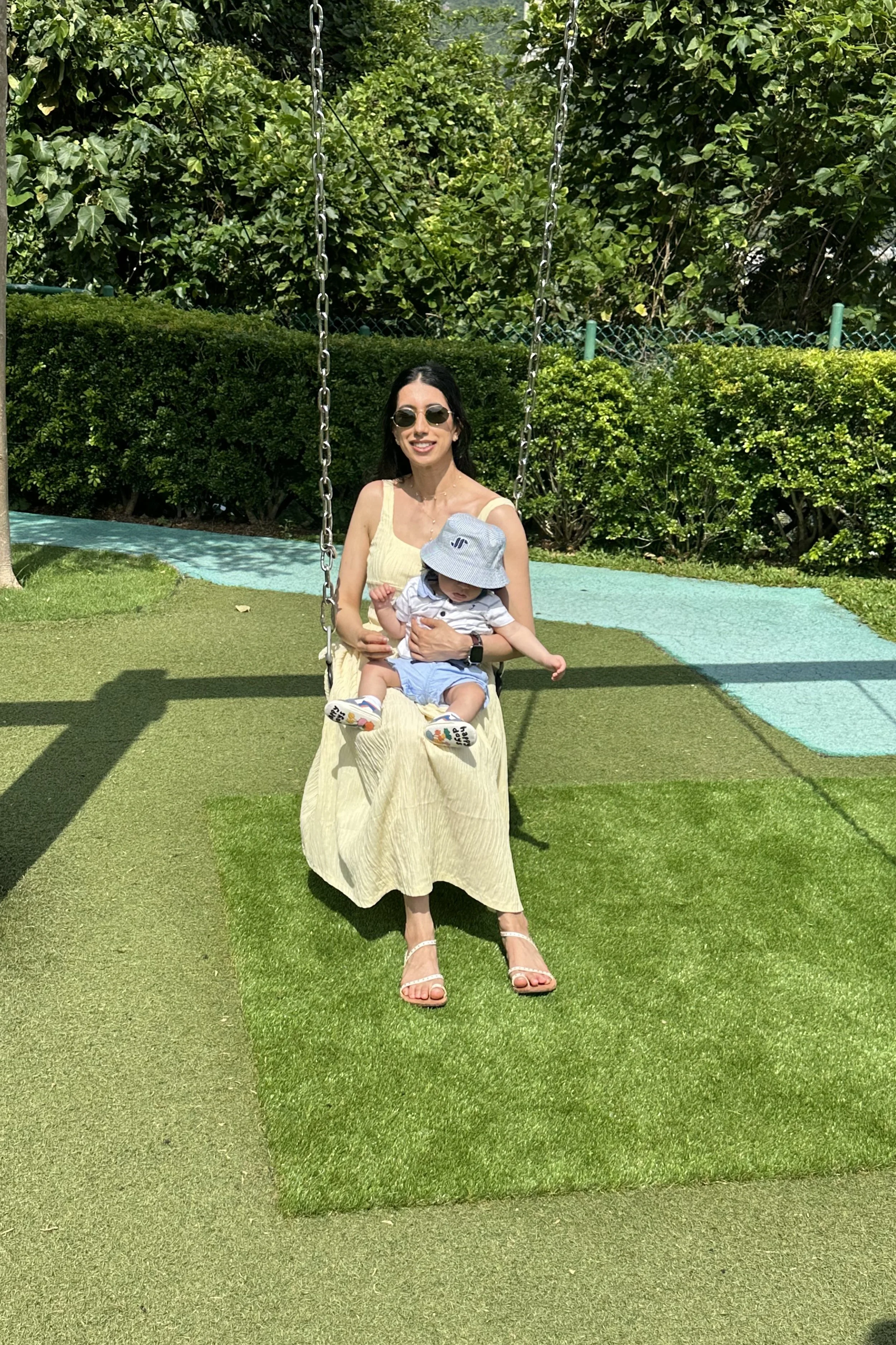 A woman sitting on a swing with a small child on her lap in a park, surrounded by green bushes and trees, with shadows cast on the grass.