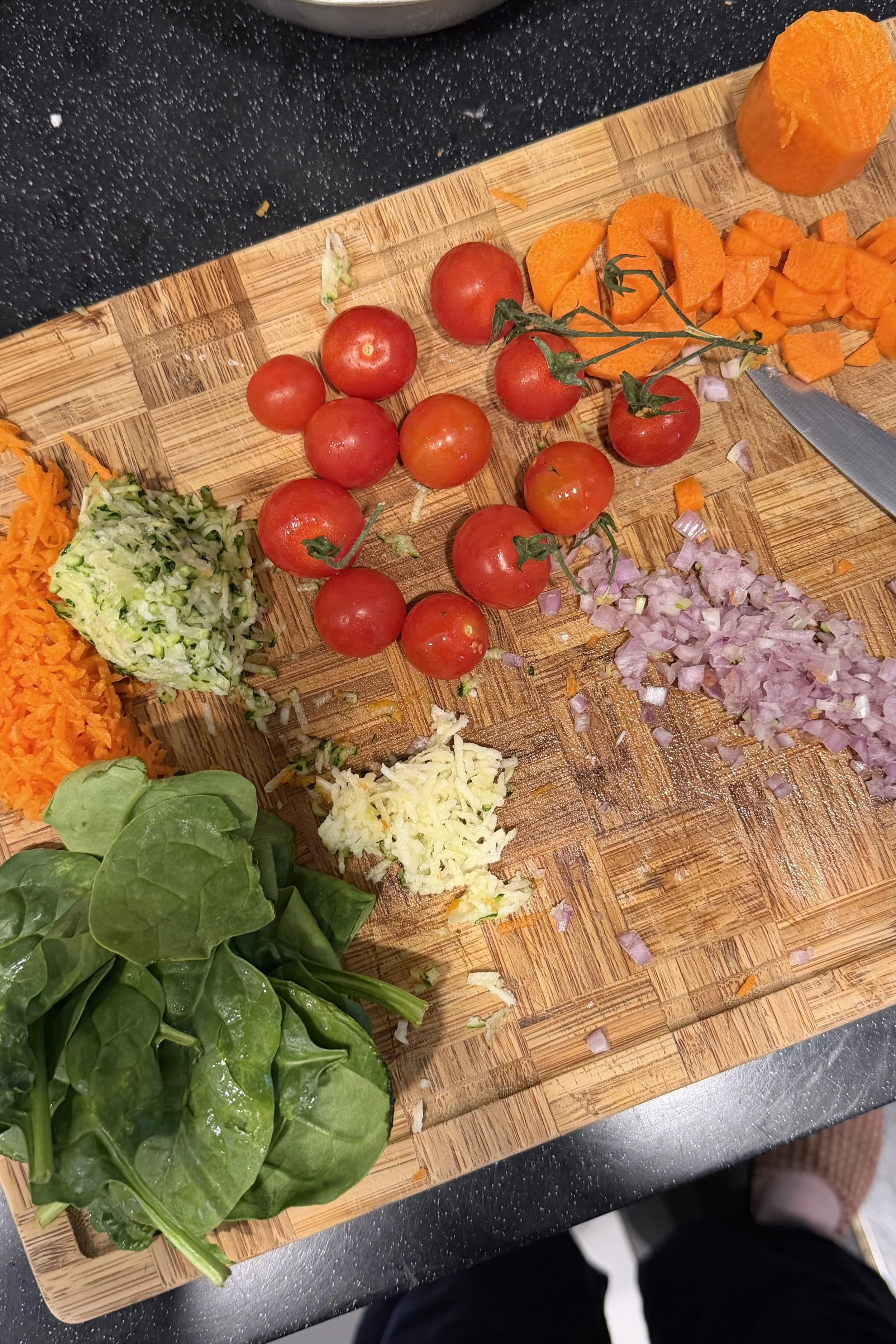 Cherry tomatoes, chopped carrots, chopped onions, chopped garlic, and fresh spinach on a wooden cutting board.