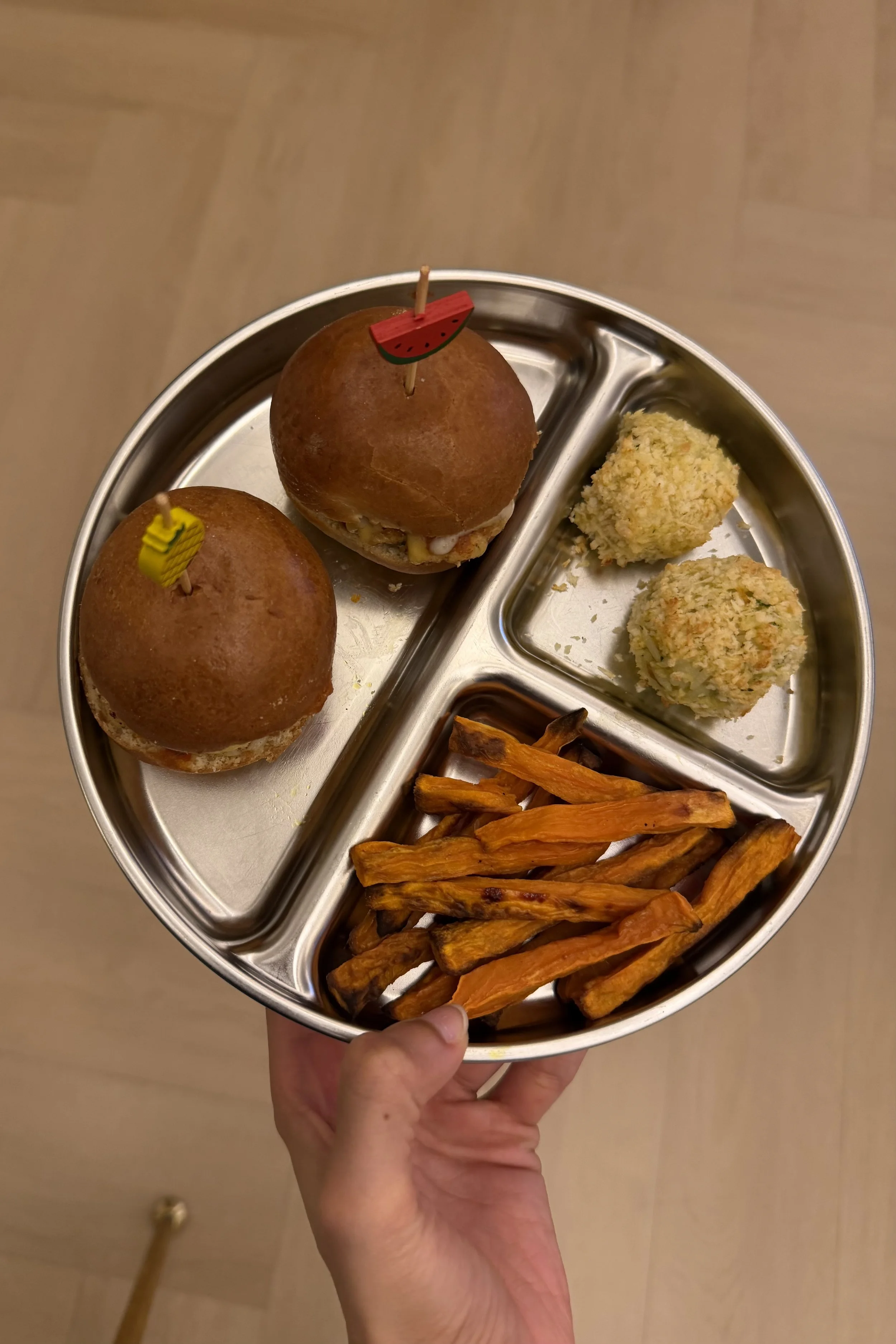 Hand holding a stainless steel divided tray with two bread rolls with fruit flags, two cheese balls, and sweet potato fries.