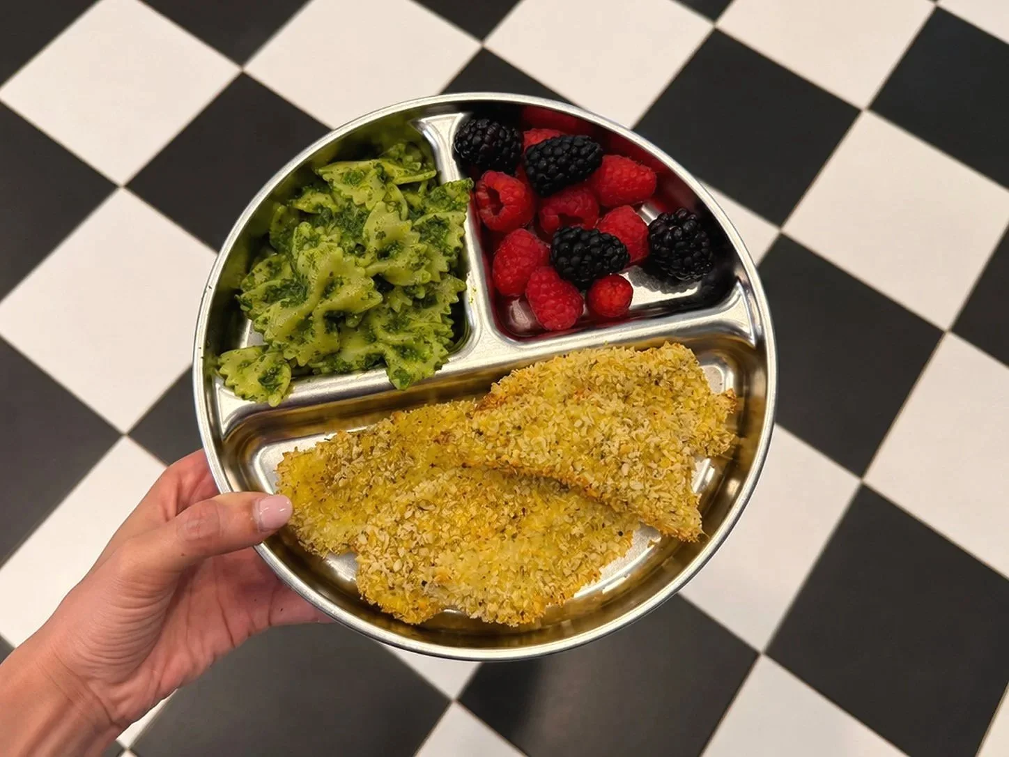 Hand holding a metal lunch tray with three compartments containing broccoli, mixed berries, and breaded fish fillets, against black and white checkered floor.