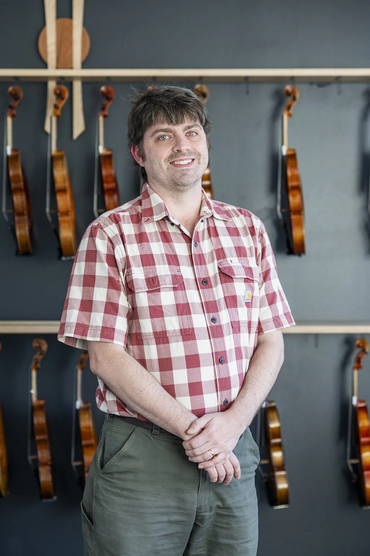 A man in a red and white checkered shirt standing in front of a wall with multiple guitars hanging on it.