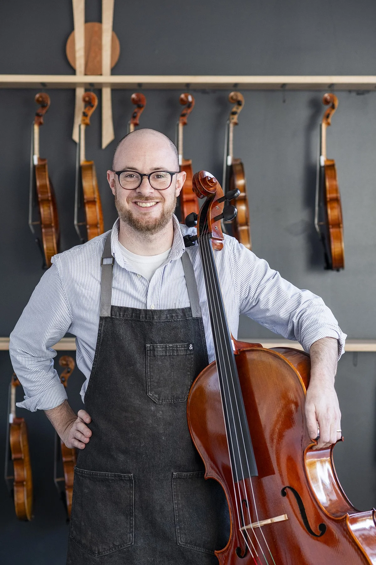 A man with glasses and a beard smiling while holding a double bass, standing in front of a wall with several violins hanging.