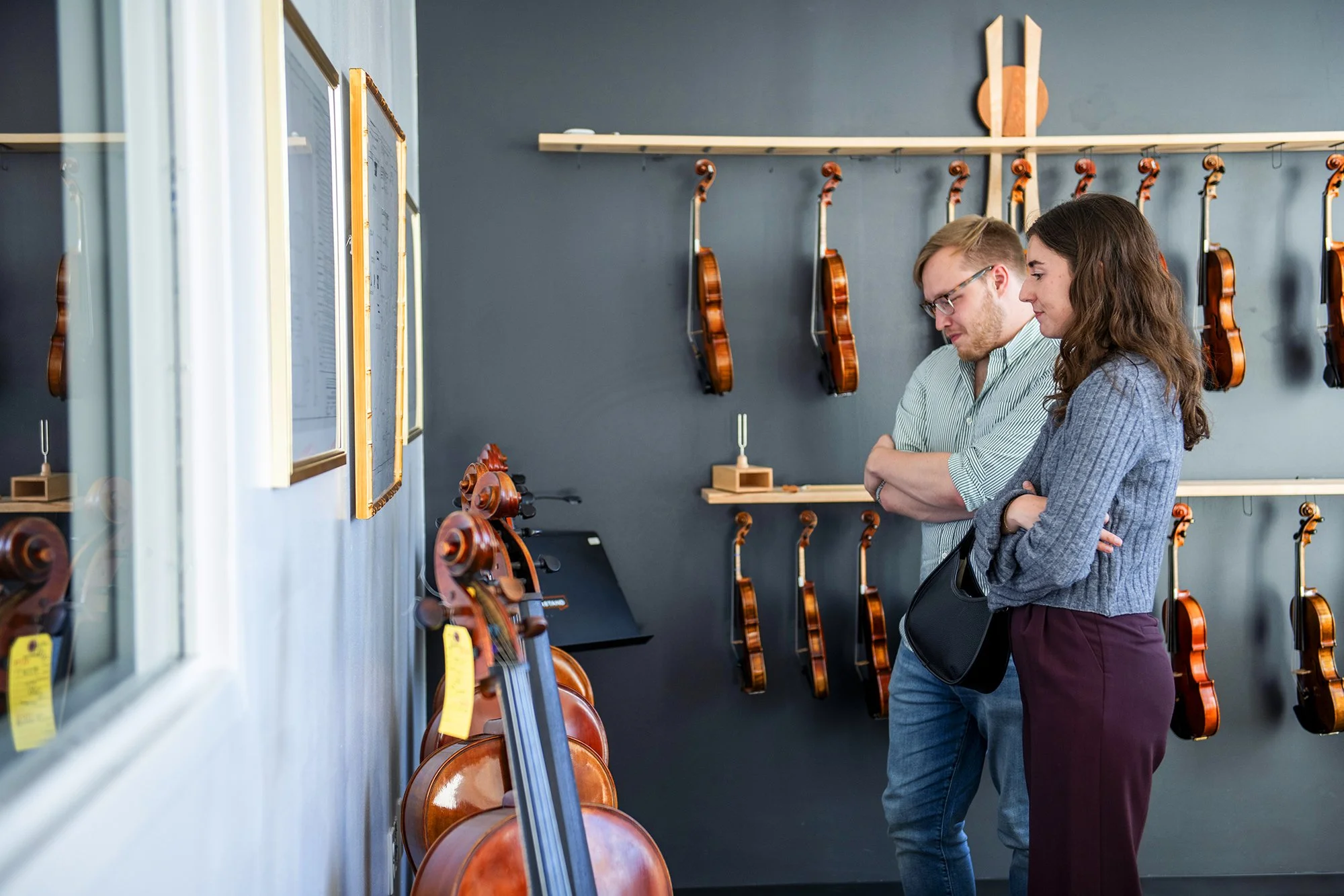 Two people, a man with glasses and a woman, are standing in a room with numerous violins hanging on the wall behind them. They are looking at items on display, possibly in a music store or exhibit.