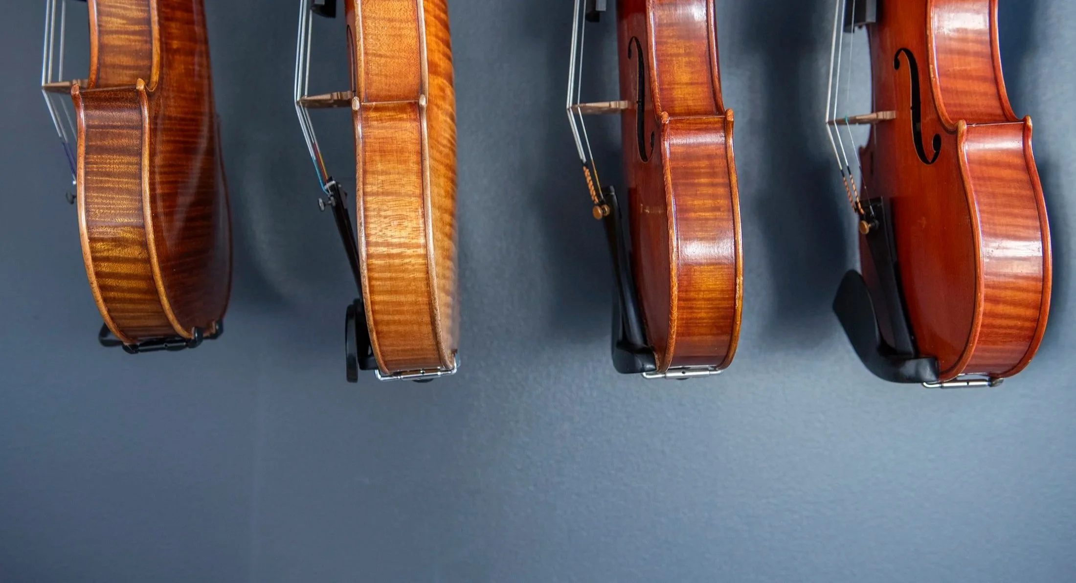 Four violins with similar wood finish are displayed on black stands, arranged in a row against a dark background.