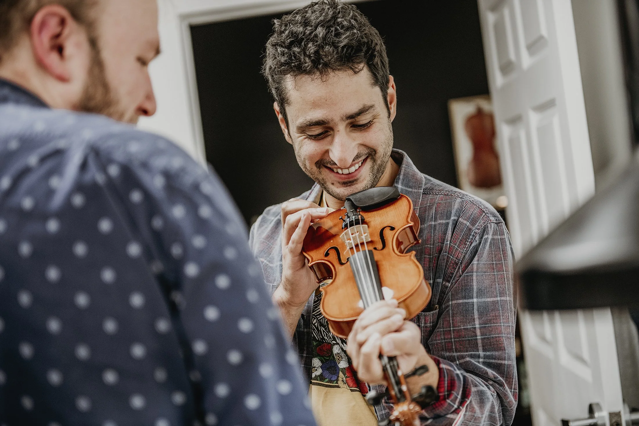 Two young men, one with dark curly hair and one with blond hair, are smiling and playing a violin together indoors, in a casual setting.