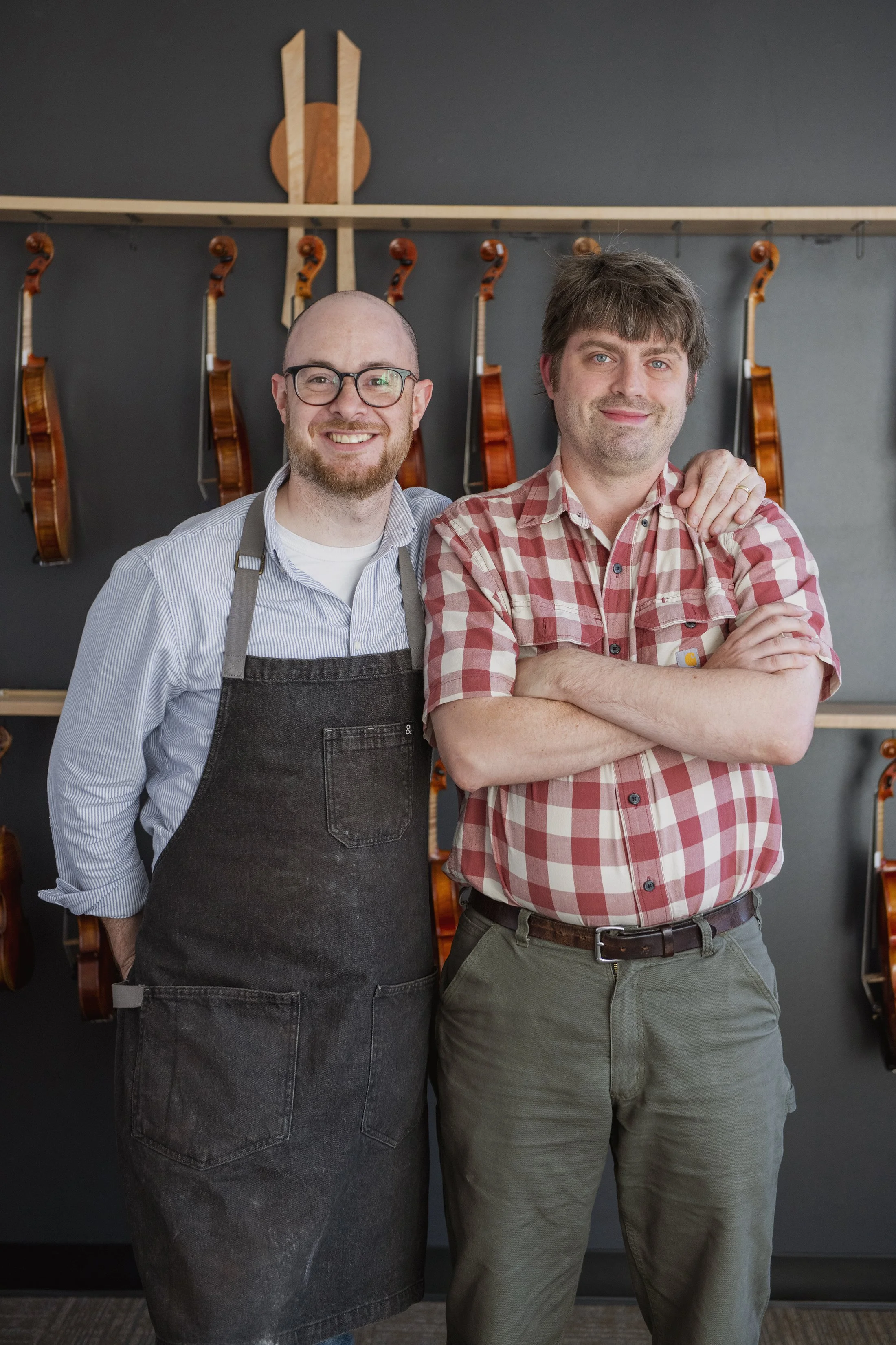 two luthiers standing with hanging violins. One man is wearing glasses, a striped shirt, and a black apron. The other man is wearing a red plaid shirt with his arms crossed. They are smiling and one has his hand on the other's shoulder.