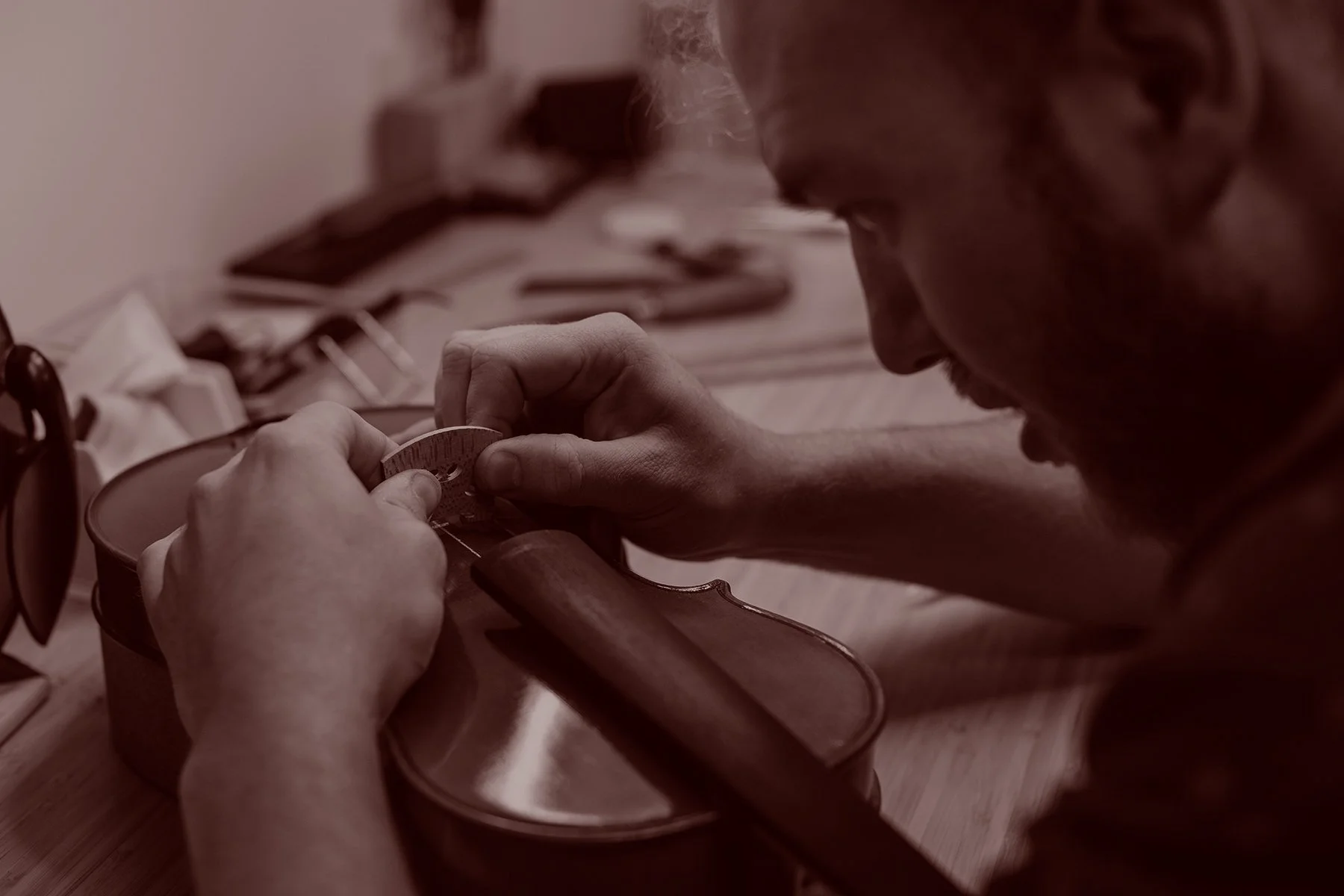 a luthier's hands working on a wooden violin, using a small tool to adjust or carve it with his hands. A wooden table with various woodworking tools can be seen in the background.
