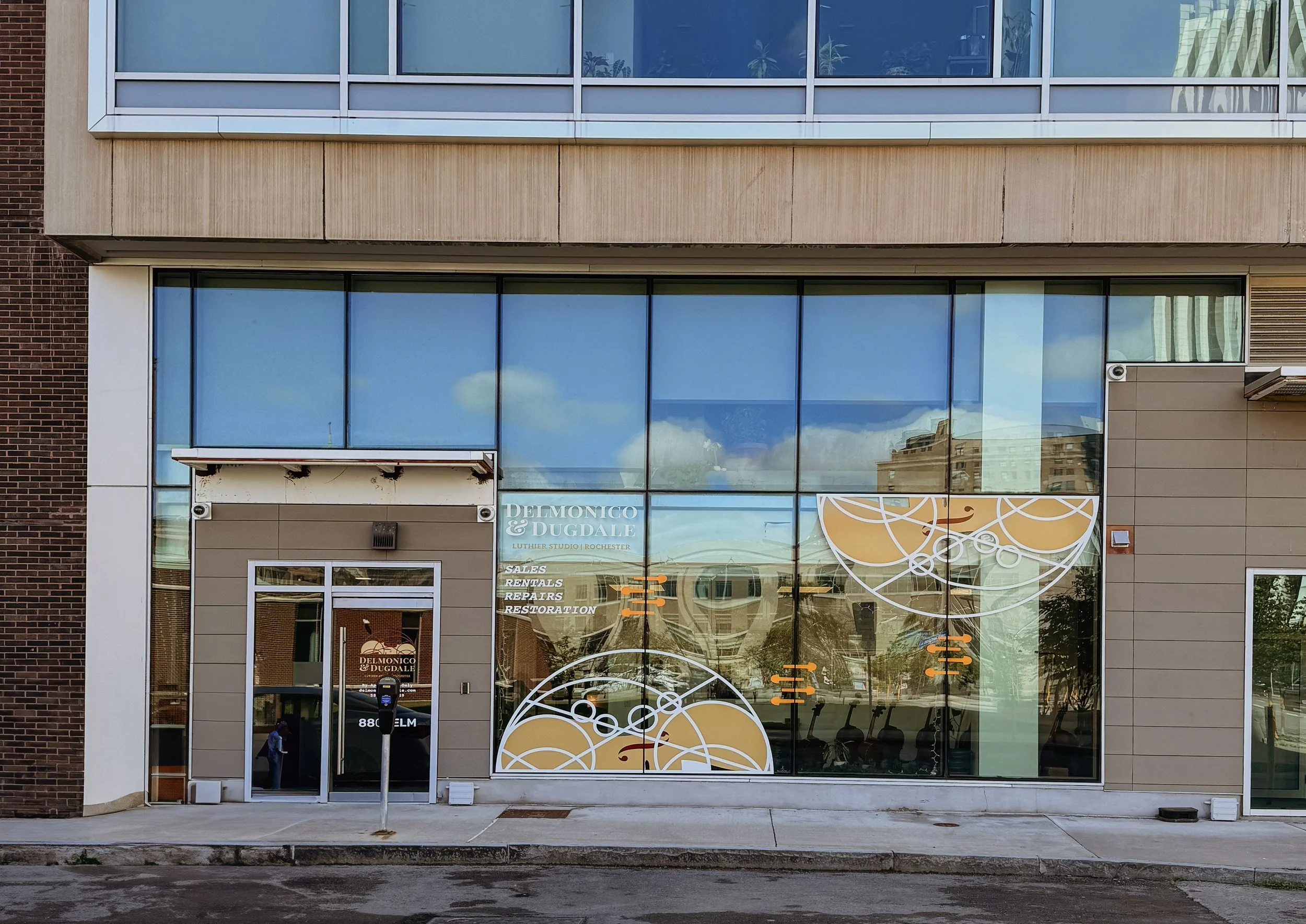 Front view of a modern commercial building with large glass windows and a sign for Delmonico & Dugdale Luthier Studio in Rochester. The building has a brick section on the left, beige siding, and decorative yellow and white graphics on the windows. There are people and chairs reflected in the glass.