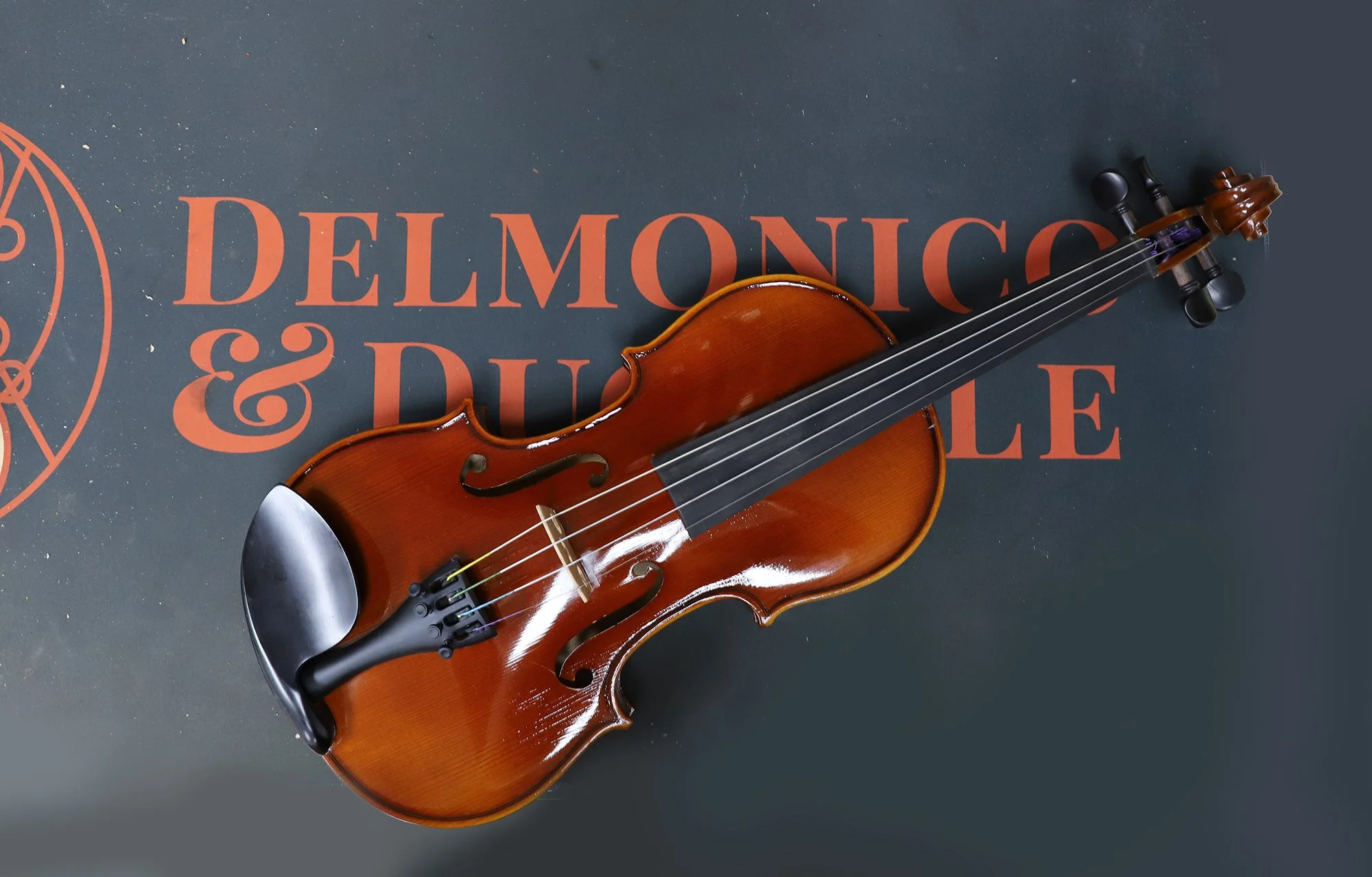 A violin resting on a dark surface with orange text that reads 'DEMONC & DUEL' and a faded geometric logo on the left.
