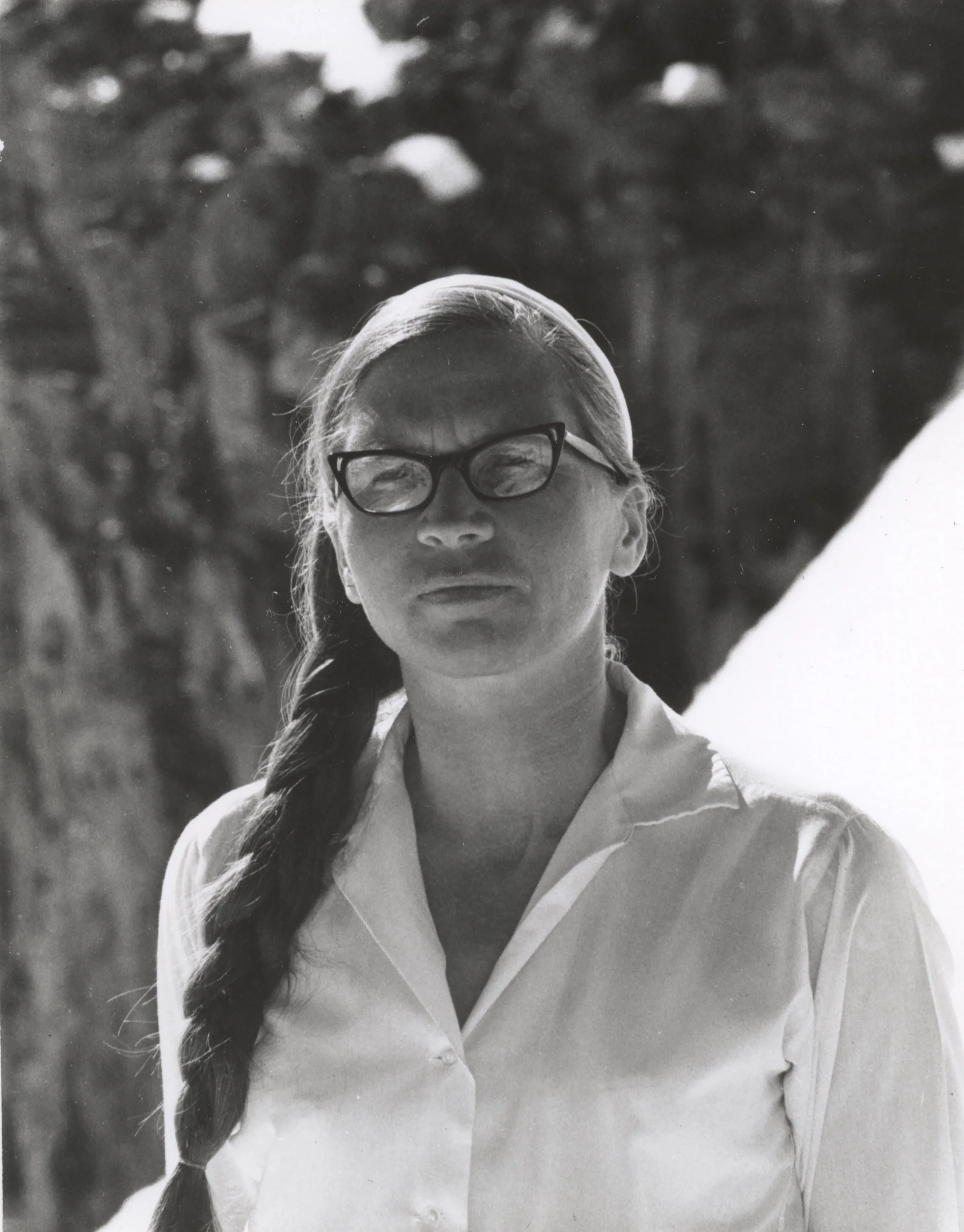 Black and white photo of a woman with long hair in a braid, wearing glasses and a collared shirt, outdoors with trees in the background.