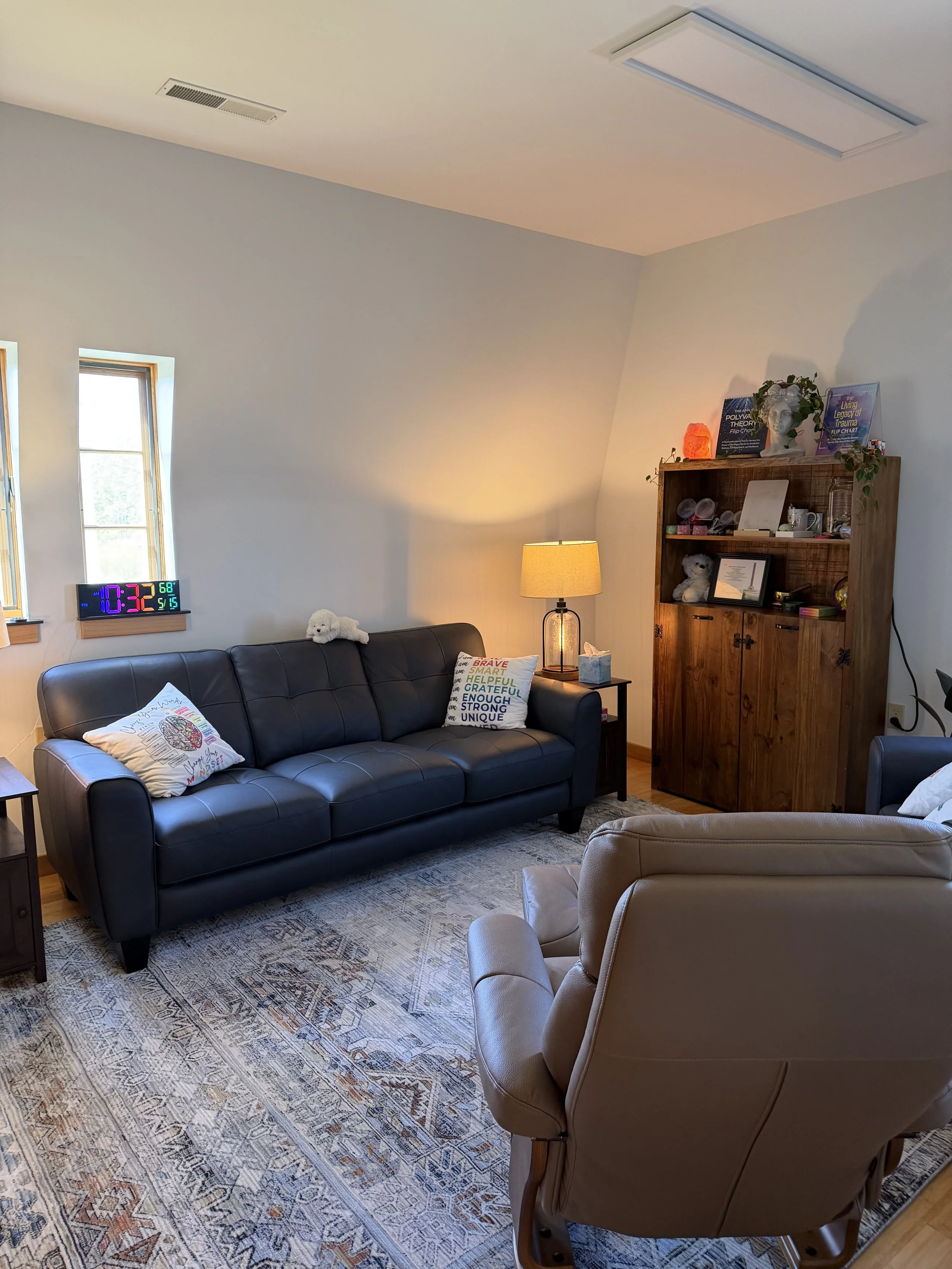 Living room with two windows, a black leather sofa with a stuffed animal and decorative pillows, a wooden bookshelf with books and decorations, a table lamp, and a tan armchair on an ornate rug.
