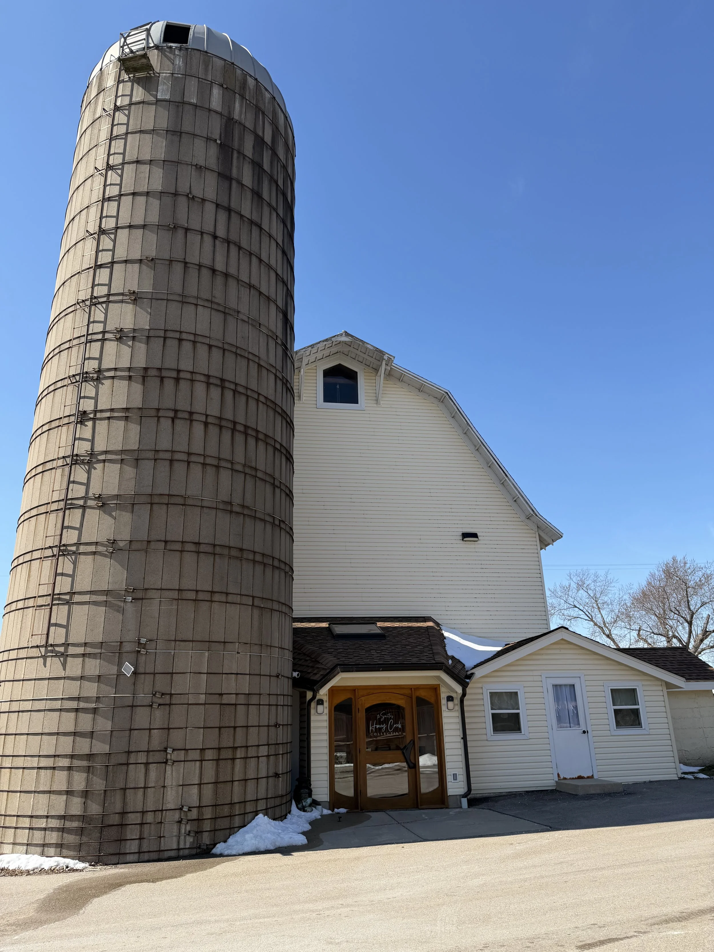 A light-colored building with a wooden entrance door and two windows, next to a large, weathered grain silo, under a clear blue sky with some trees in the background and snow on the ground.