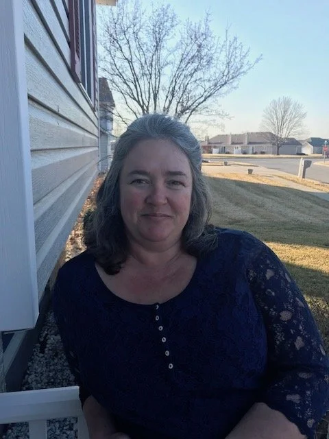 A woman with shoulder-length gray hair, wearing a dark blue lace top, sitting outside near a house with vinyl siding, with a tree and neighborhood houses in the background on a sunny day.