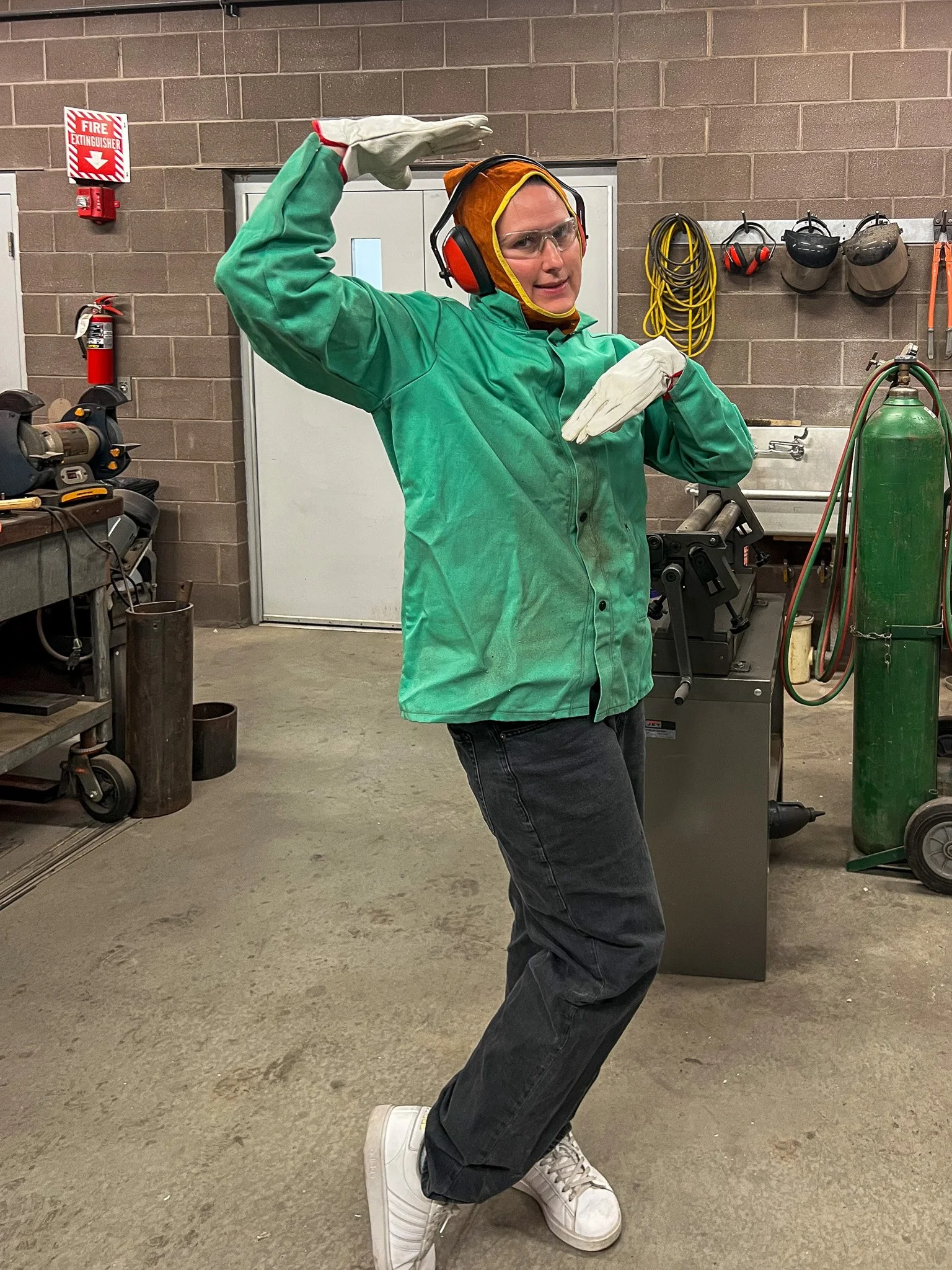 A person posing in a workshop, wearing protective gear including a green coverall, gloves, safety goggles, and a helmet with hearing protection. They have one arm raised above their head and the other arm across their chest.