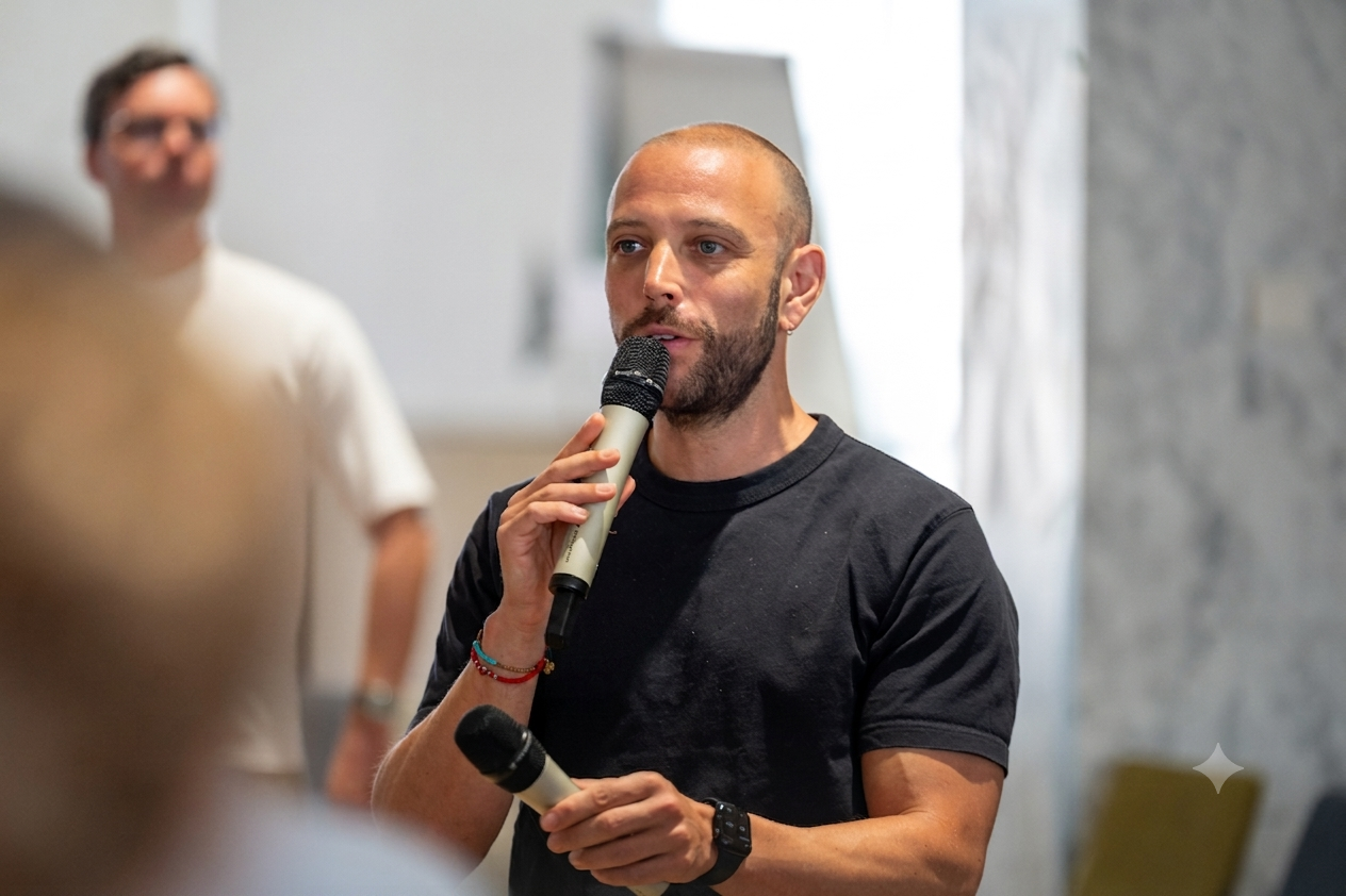 A man with a beard and short hair speaking into a handheld microphone during an indoor event, wearing a black T-shirt, with a blurred background and an audience member in the foreground.
