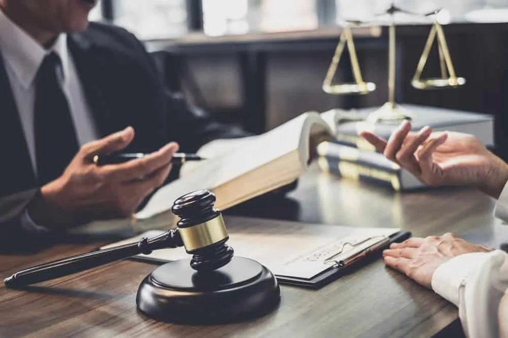 Two people in a legal office discussing, with a gavel, books, a scale of justice, and documents on the table.