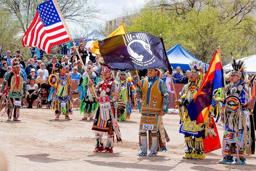 Native American dancers in traditional costumes and headdresses, holding flags including the U.S. flag and a tribal flag, performing at a powwow or cultural event with spectators in the background.