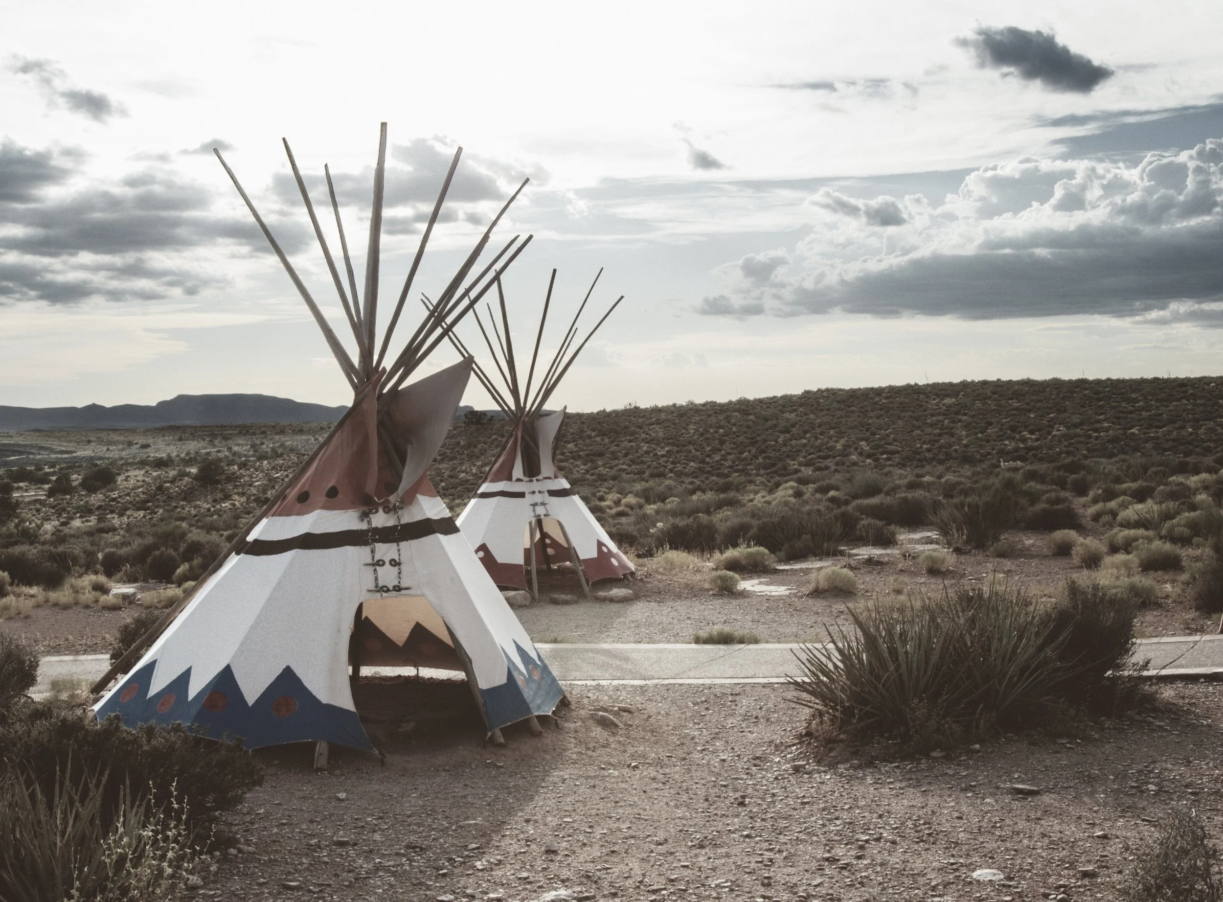 Row of tipi tents with painted designs in a desert landscape, with hills in the background and a cloudy sky overhead.