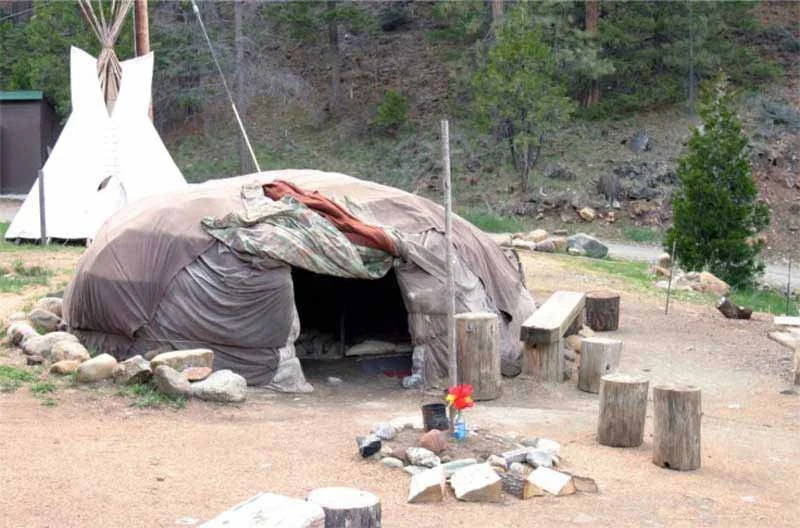 A small, tent-like structure made of fabric and logs, with a makeshift entrance, surrounded by rocks and outdoor furniture, in a natural setting with trees and hills in the background.