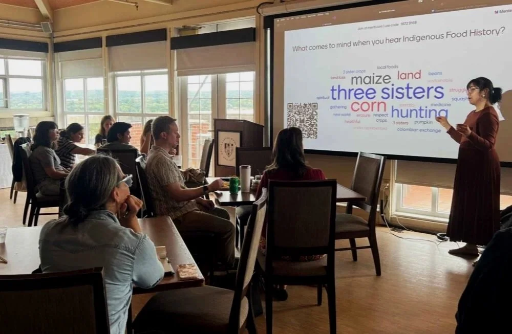 A woman in a brown dress giving a presentation to a group of people in a room with large windows, with a slide on the screen showing words related to Indigenous Food History, including 'three sisters,' 'corn,' 'hunting,' and 'land.'