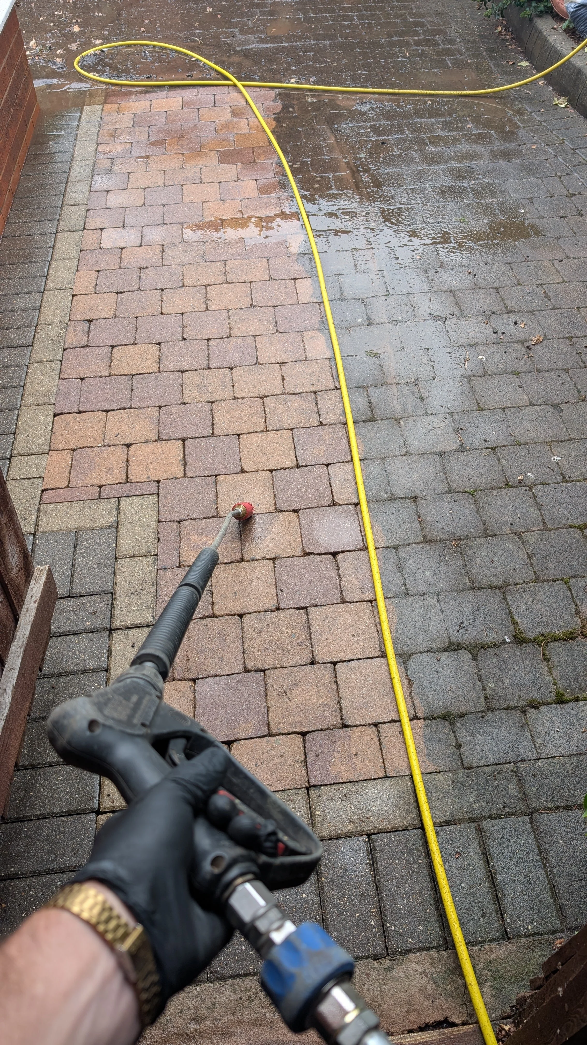 A person wearing a black glove is using a pressure washer to clean a brick patio, with a yellow hose running across wet bricks.