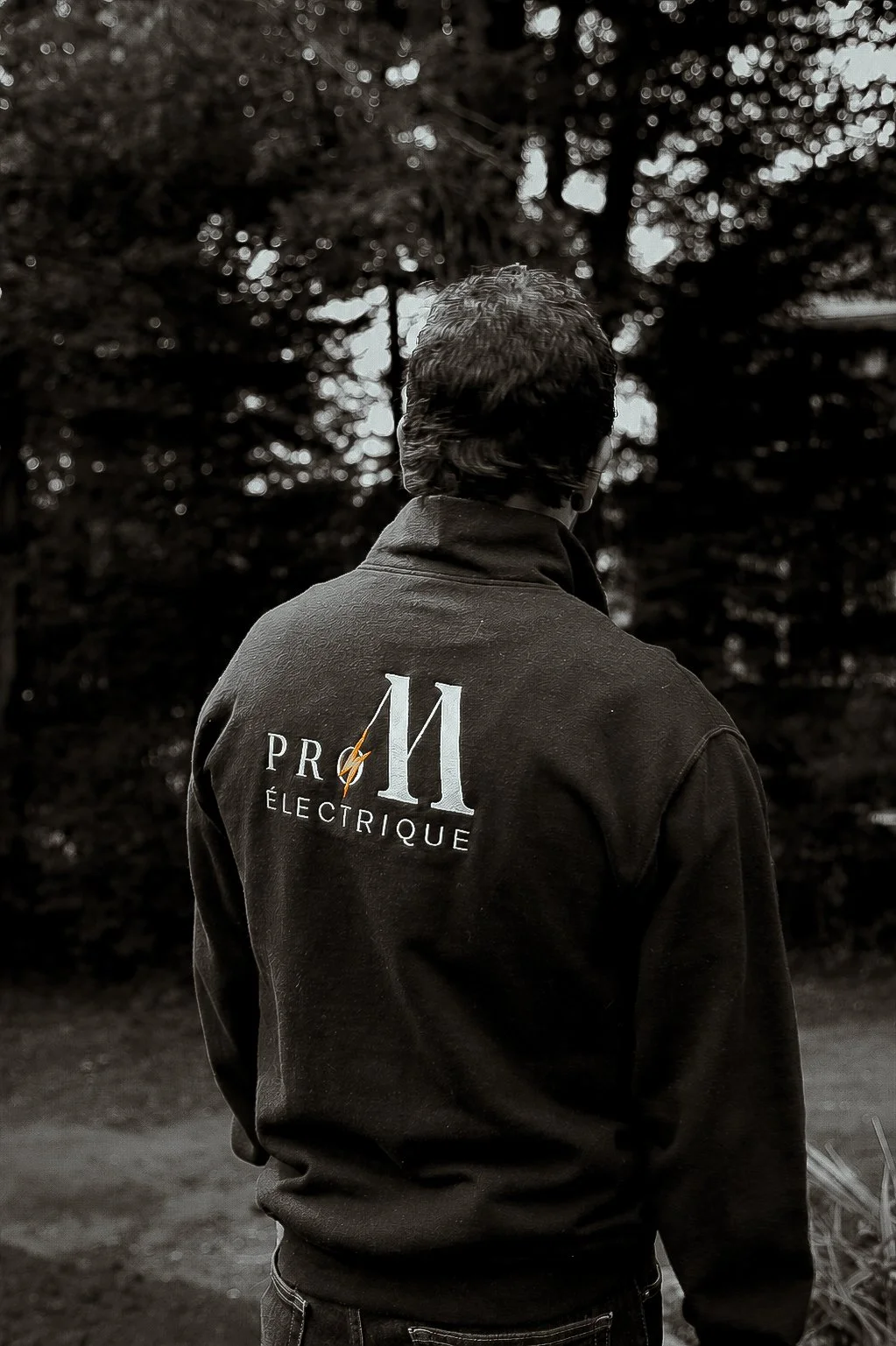 Une personne de dos portant une veste noire avec le logo 'Pro 11 Électrique' dans un environnement naturel avec des arbres.