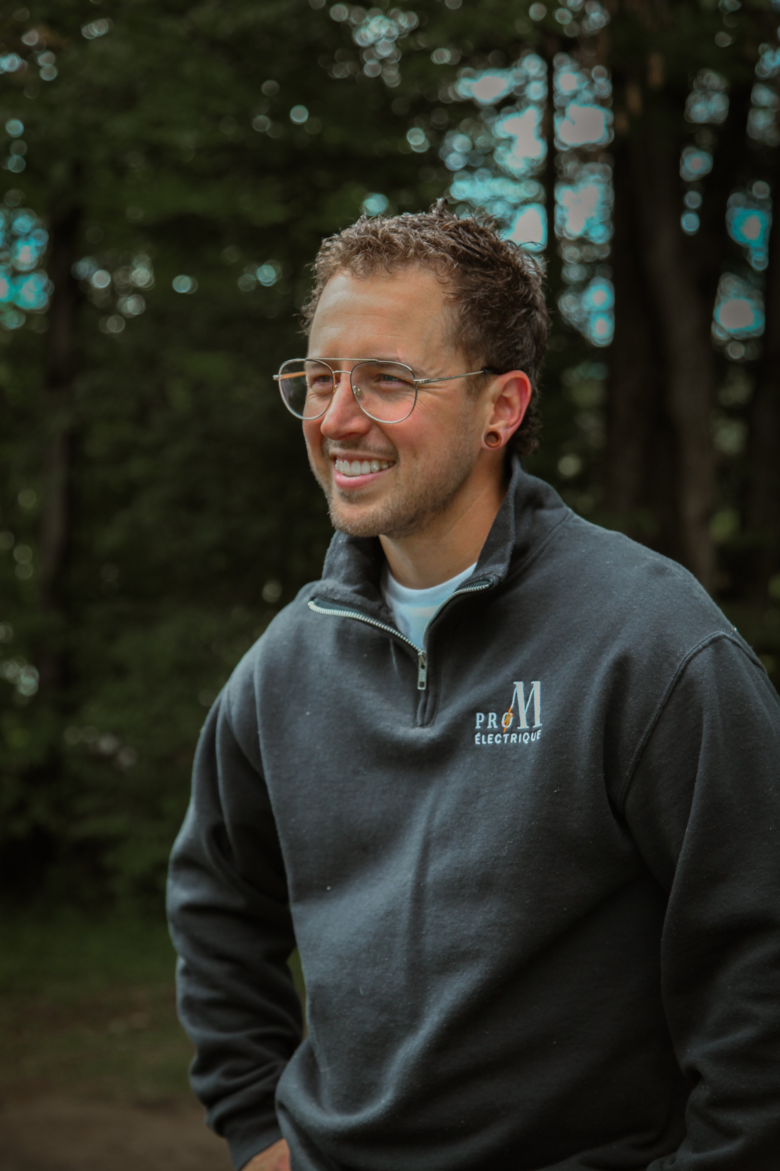 Un homme souriant portant des lunettes, une veste gris foncé avec un logo 'PR O M É lectrique' sur fond d'arbres.