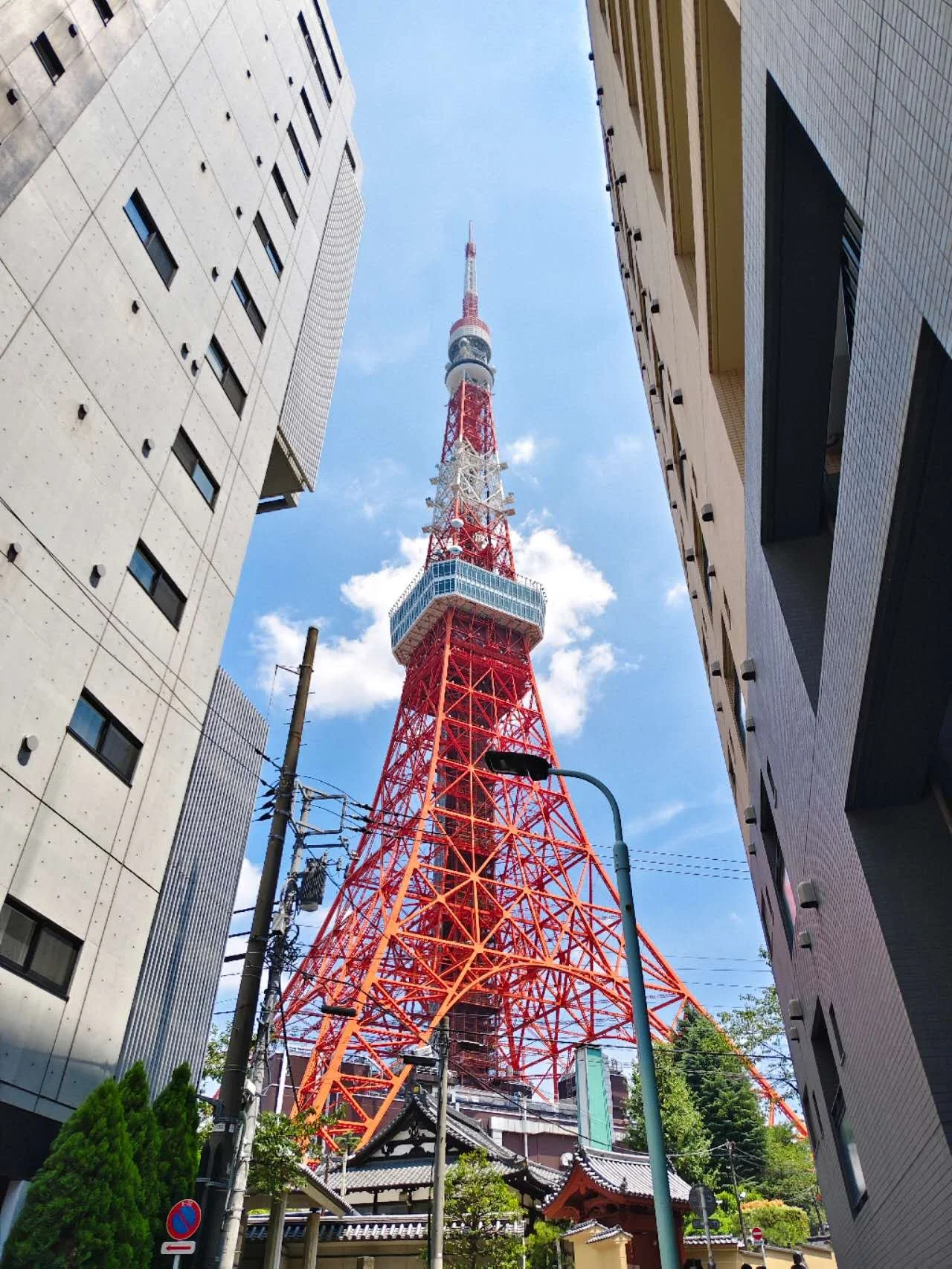 Tokyo Tower framed by modern buildings and traditional architecture with trees and street signs in the foreground, under a blue sky with some clouds.