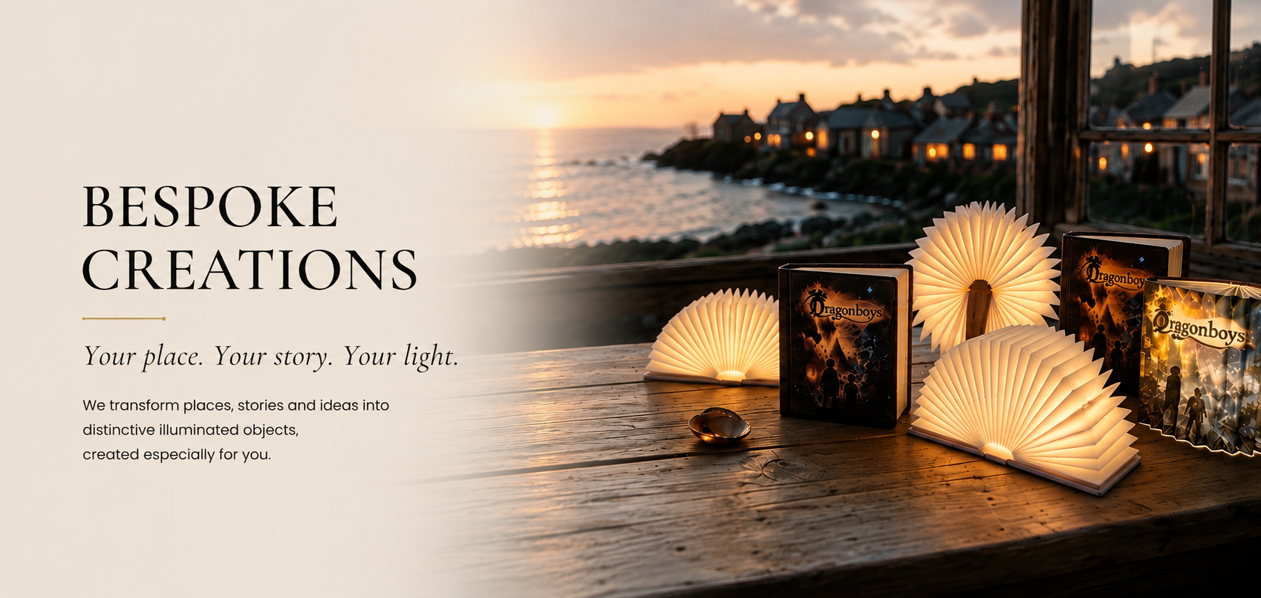 A promotional image with a sunset view over a coastal village, featuring illuminated decorative objects, including book-shaped lights and fan-shaped lamps, on a wooden surface with a window in the background.