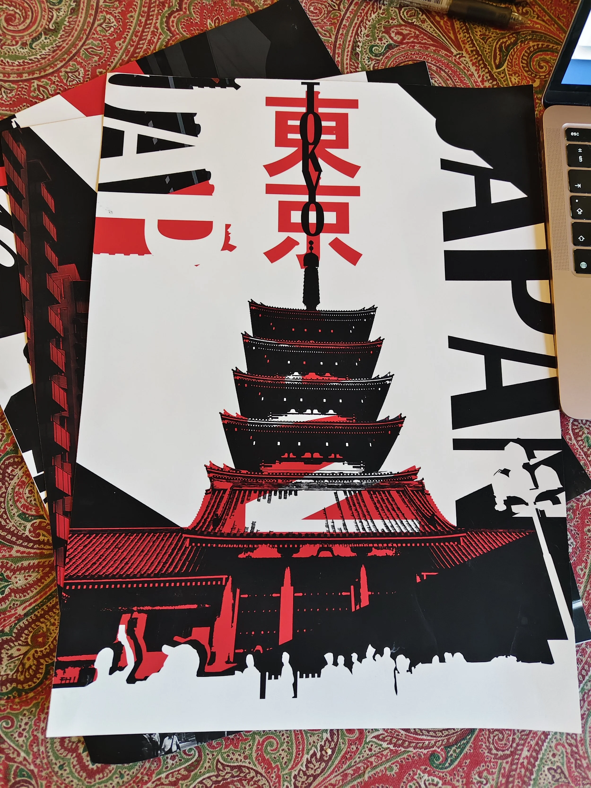 Black, white, and red poster of the Tokyo Tower with silhouettes of people at the bottom, displayed on a patterned tablecloth.
