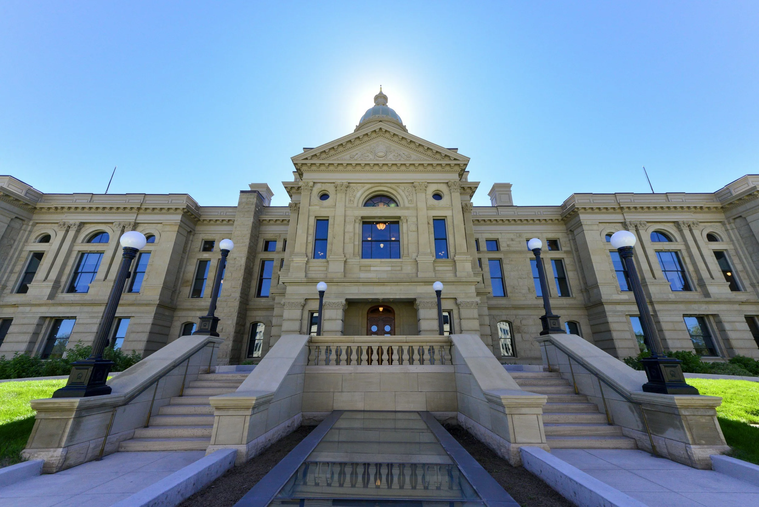 Front view of a historic courthouse with stairs, columns, and a clock, under a clear blue sky with the sun behind the roof.