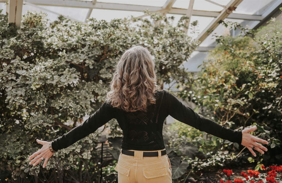 A woman with curly hair wearing a black long-sleeve shirt and beige pants stands with her arms outstretched inside a greenhouse surrounded by lush green plants and flowers.