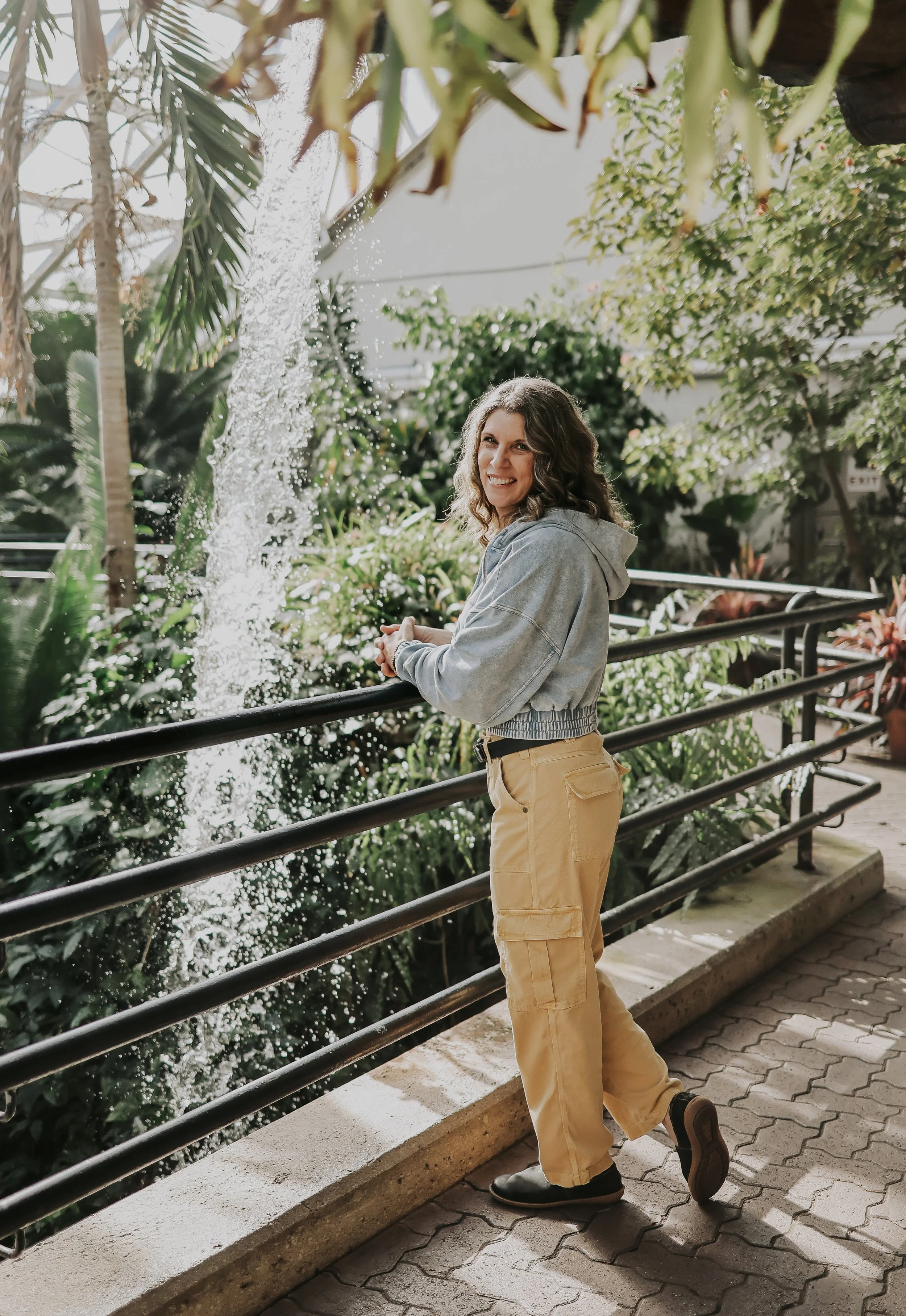 A woman with curly hair smiling while standing near a railing in a lush indoor botanical garden, wearing a gray hoodie and yellow cargo pants.