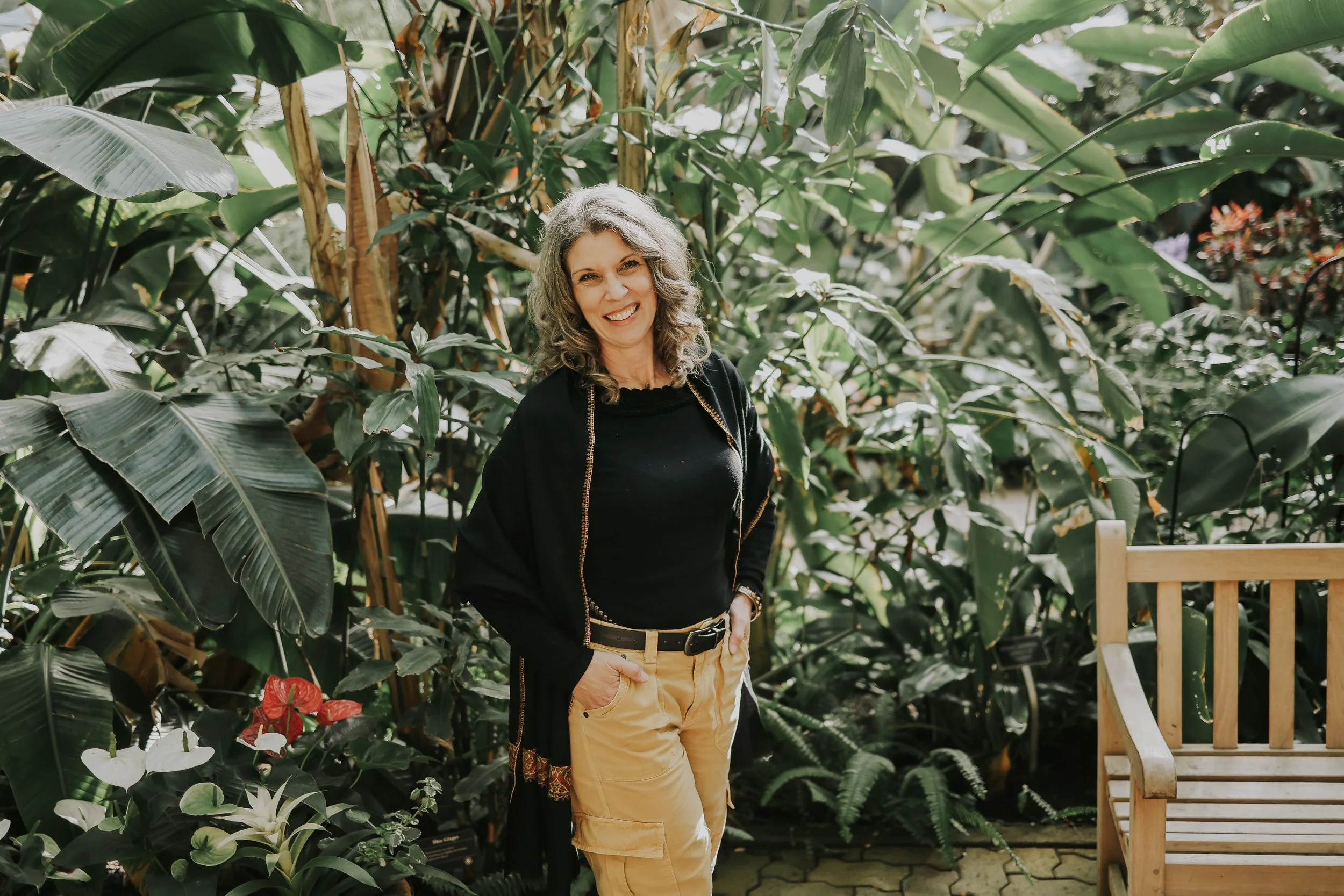 A woman with gray curly hair smiling and standing in a lush greenhouse with green foliage and colorful flowers around her.