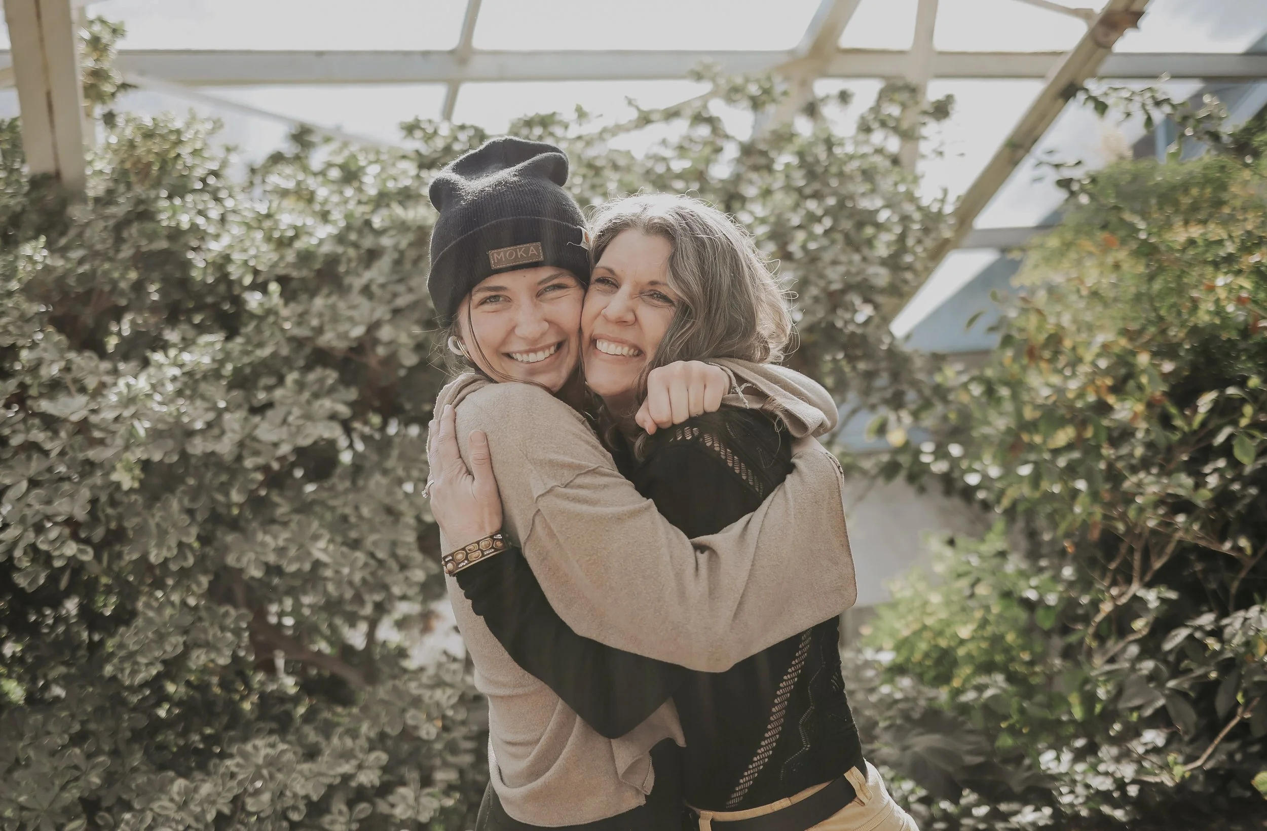 Two women hugging and smiling outdoors, one wearing a black beanie and the other with curly hair, with green foliage and a white structure in the background.