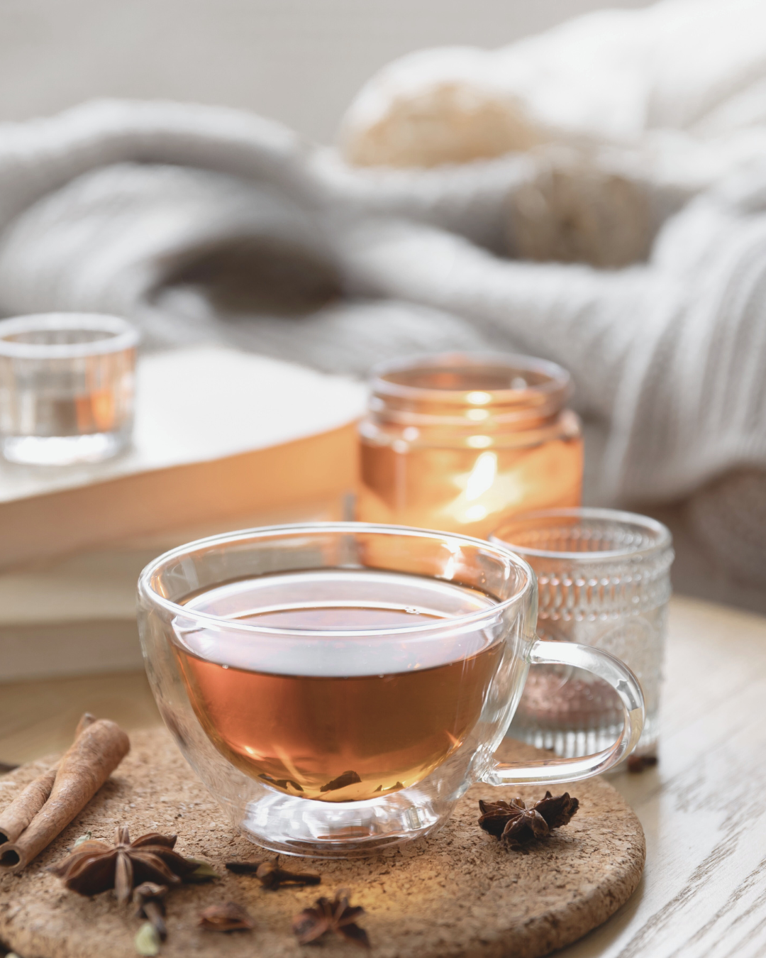 A glass cup of tea on a cork coaster, surrounded by cinnamon sticks and star anise, with candles and cozy textiles in the background.
