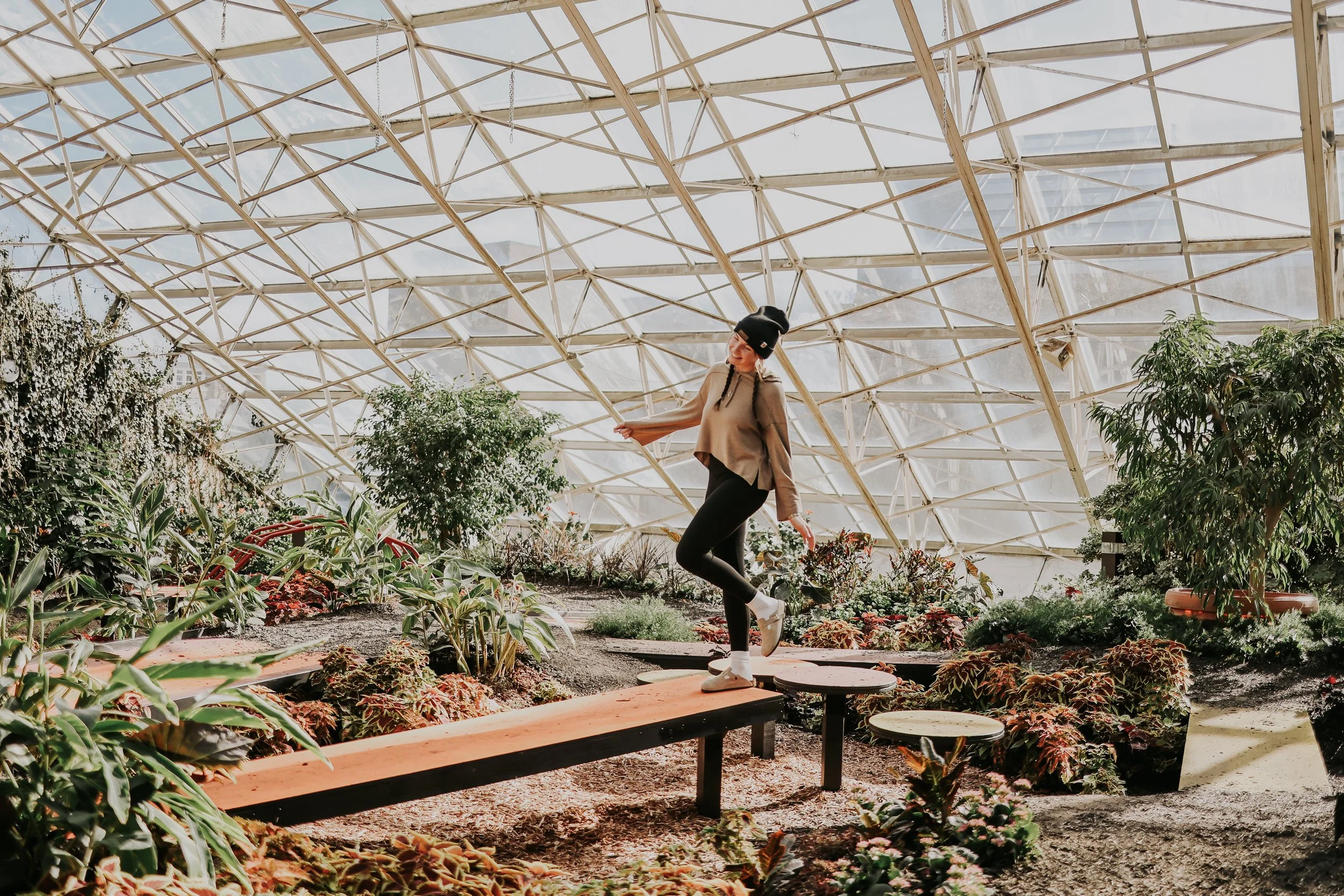 A woman in a black beanie, beige jacket, black pants, and white sneakers is posing and smiling in a greenhouse surrounded by green plants and flowers, under a curved glass and wood ceiling structure.
