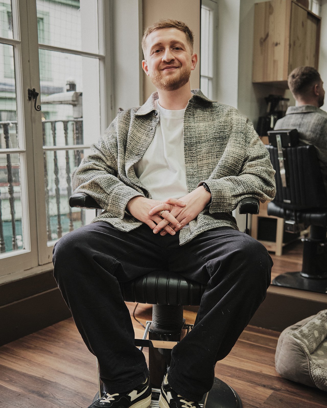 A young man with short blonde hair and a beard sitting in a barber chair inside a modern barber shop, smiling at the camera. He is wearing a white T-shirt and a gray patterned jacket, with black pants and sneakers. Large windows behind him allow natural light to fill the space.