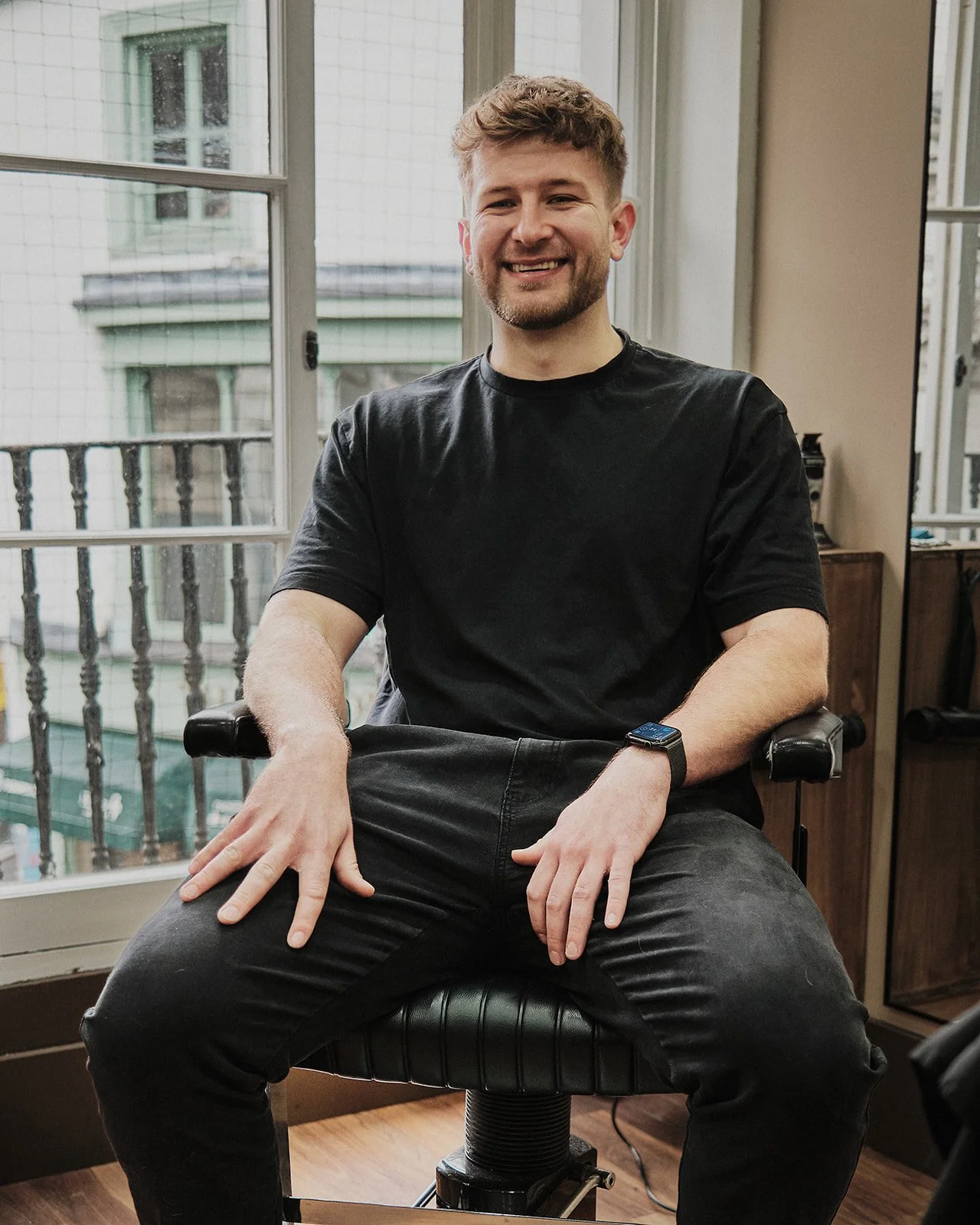 A man with short, curly hair and a beard smiling while sitting on a black chair in front of a window with a city scene outside. He is wearing a black T-shirt, black pants, and a smartwatch.
