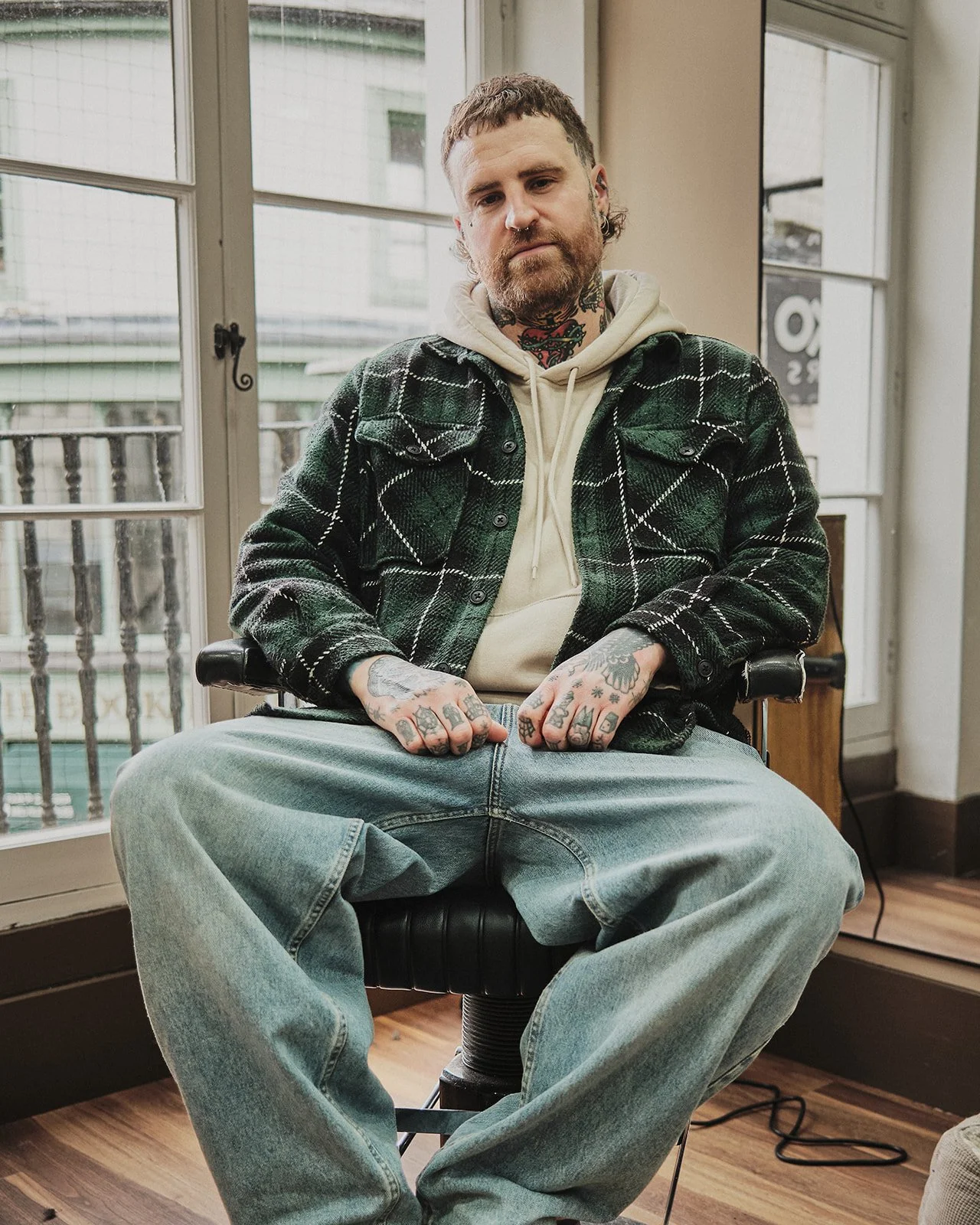A young man with tattoos and facial piercings sitting in a modern apartment, wearing a beige hoodie and a green plaid jacket, with jeans, sitting in a black office chair with large windows and a balcony in the background.